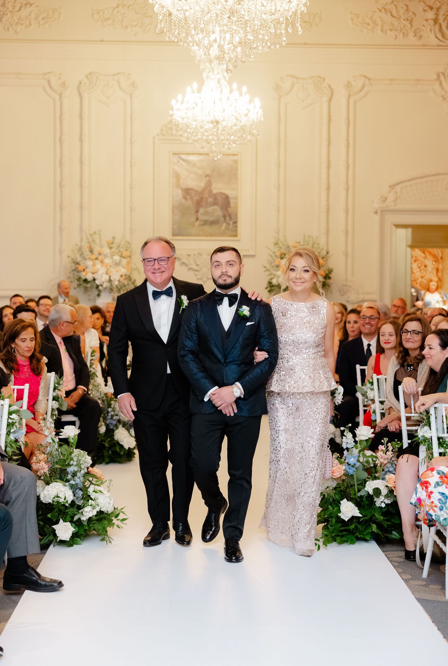 Wedding ceremony processional in an ornate ballroom with crystal chandeliers and white floral aisle decorations.