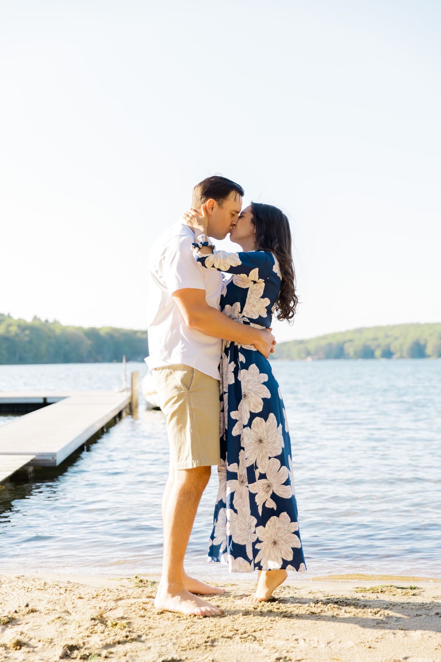 A romantic embrace on a sandy beach with a lake and wooden dock in the background.
