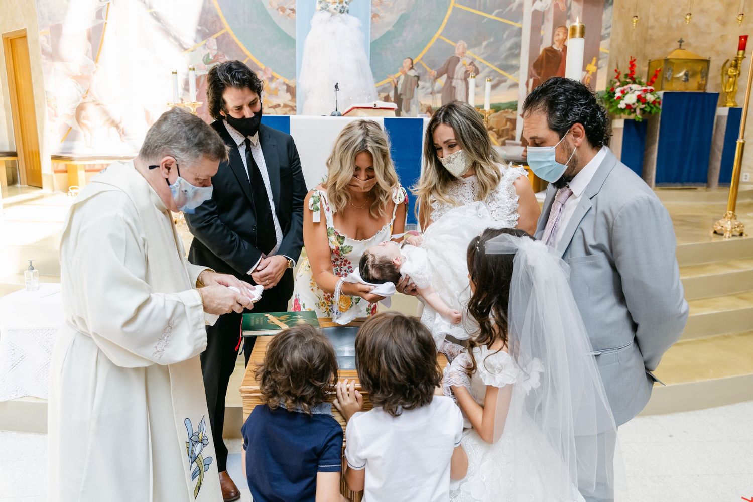Clergy and family members participate in an indoor baptism ceremony while wearing face masks.