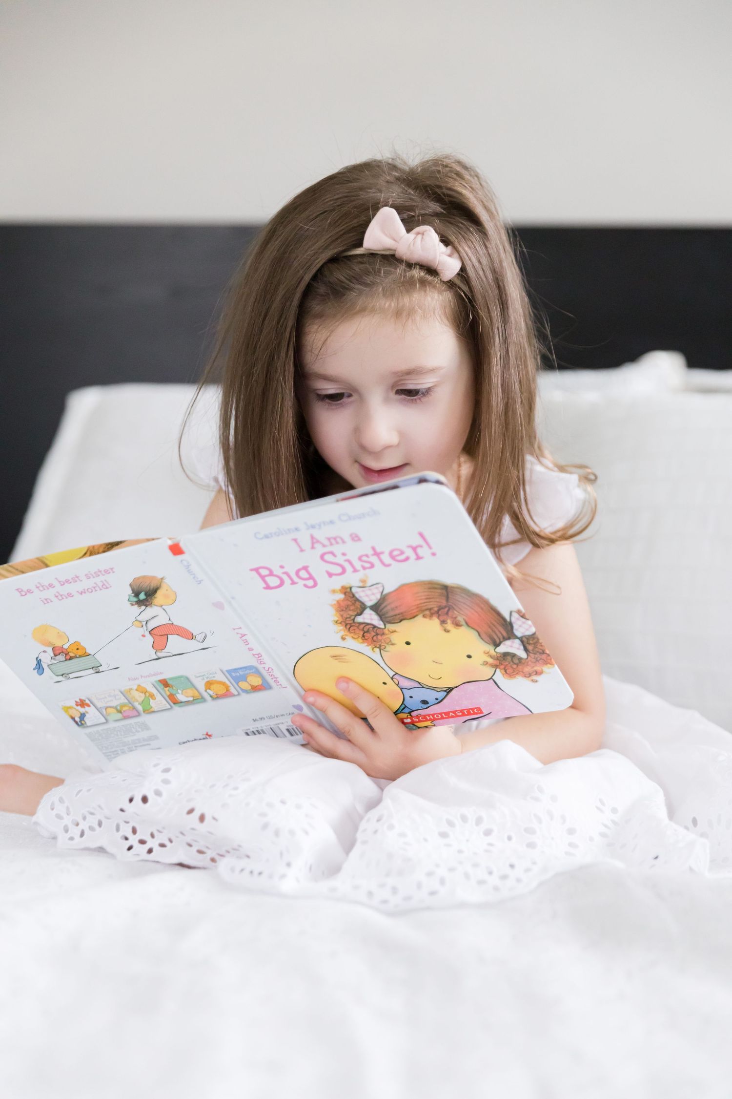 Child reading 'Big Sister' book while lying on white bedding.