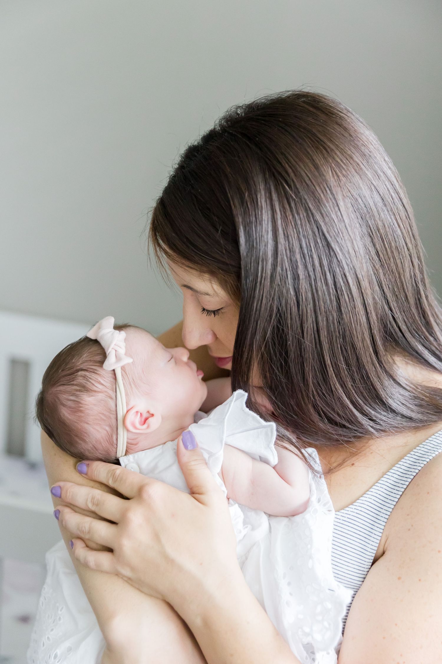 Mother in grey tank top tenderly kisses newborn in white dress.