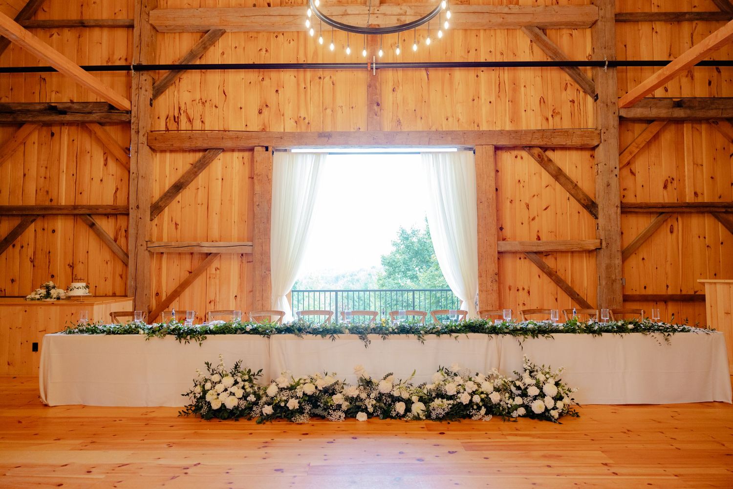 Rustic barn wedding head table with white floral decorations and draped window overlooking outdoor scenery.