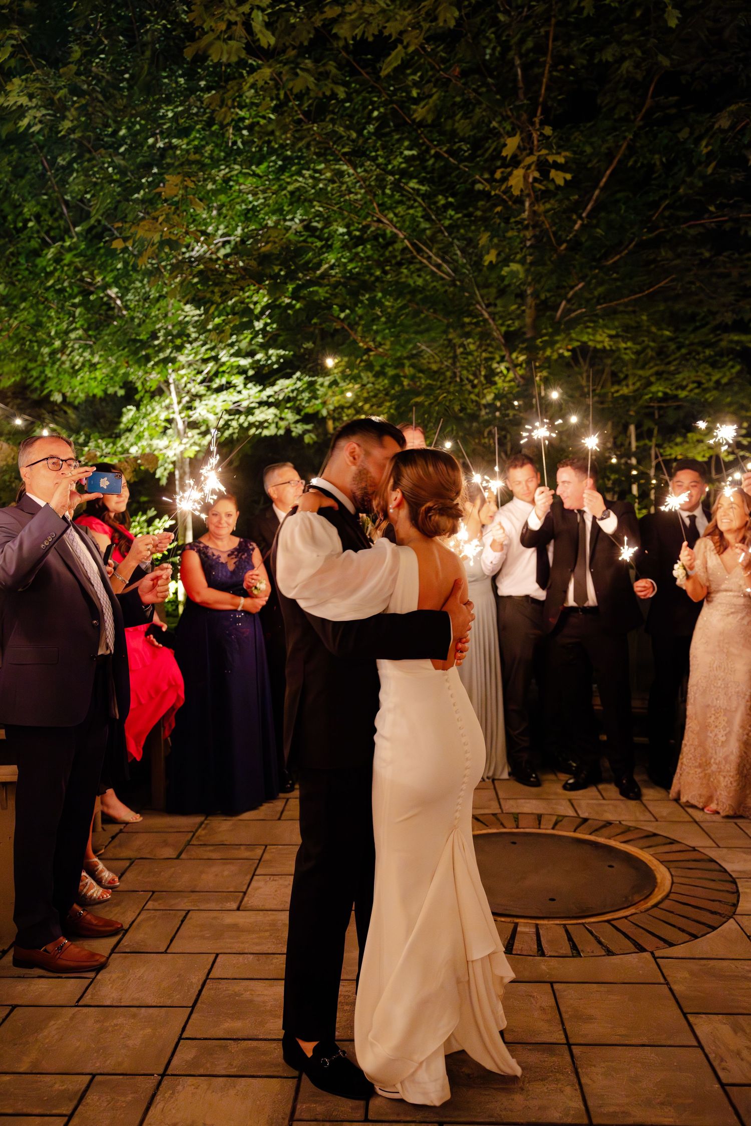 A couple shares their first dance surrounded by guests on an outdoor stone patio at night.