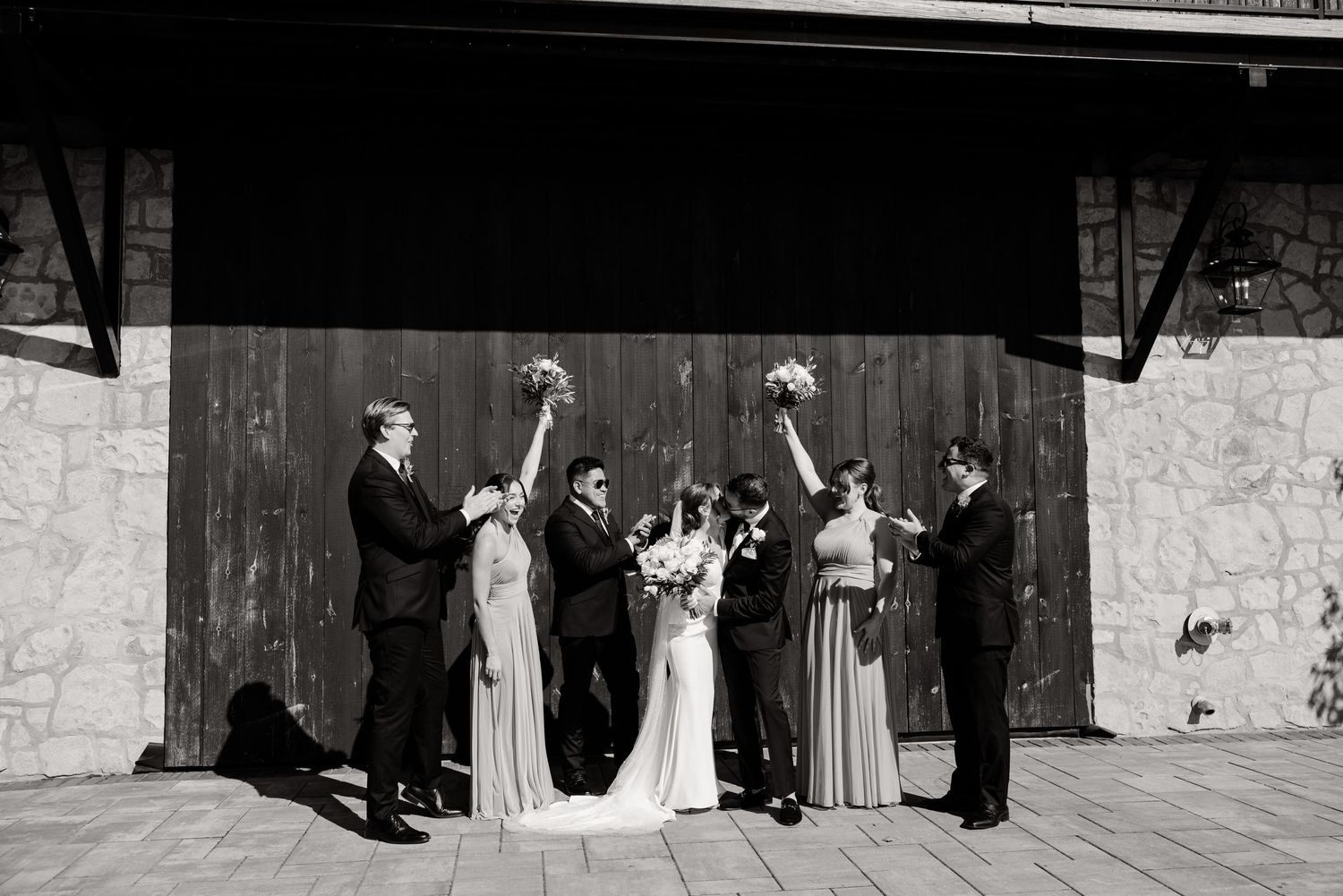 Black and white photo of wedding party celebrating with raised arms against rustic backdrop.