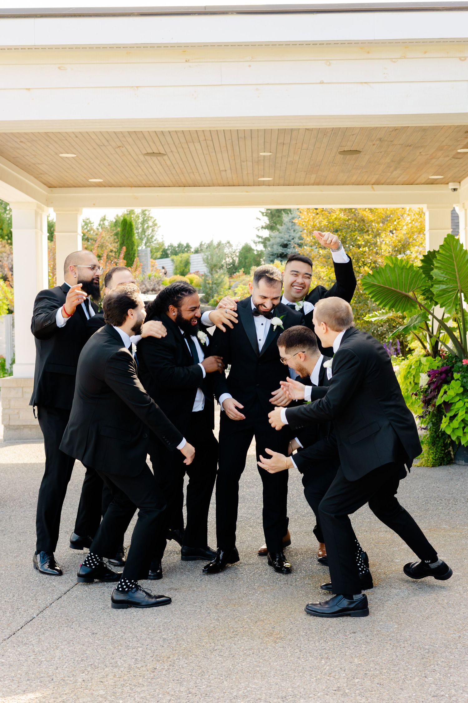 Groomsmen in black tuxedos playfully posing and celebrating together on a covered porch.
