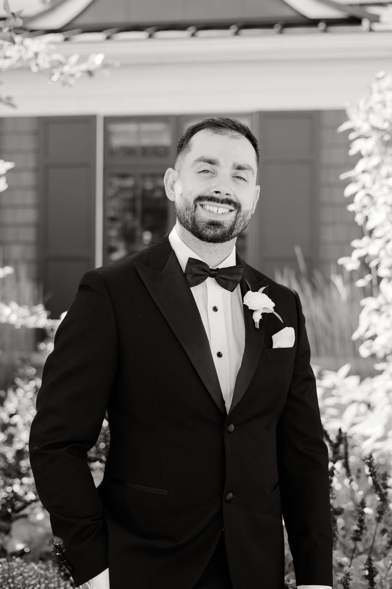 Classic black and white portrait of groom in tuxedo with boutonniere against floral backdrop.