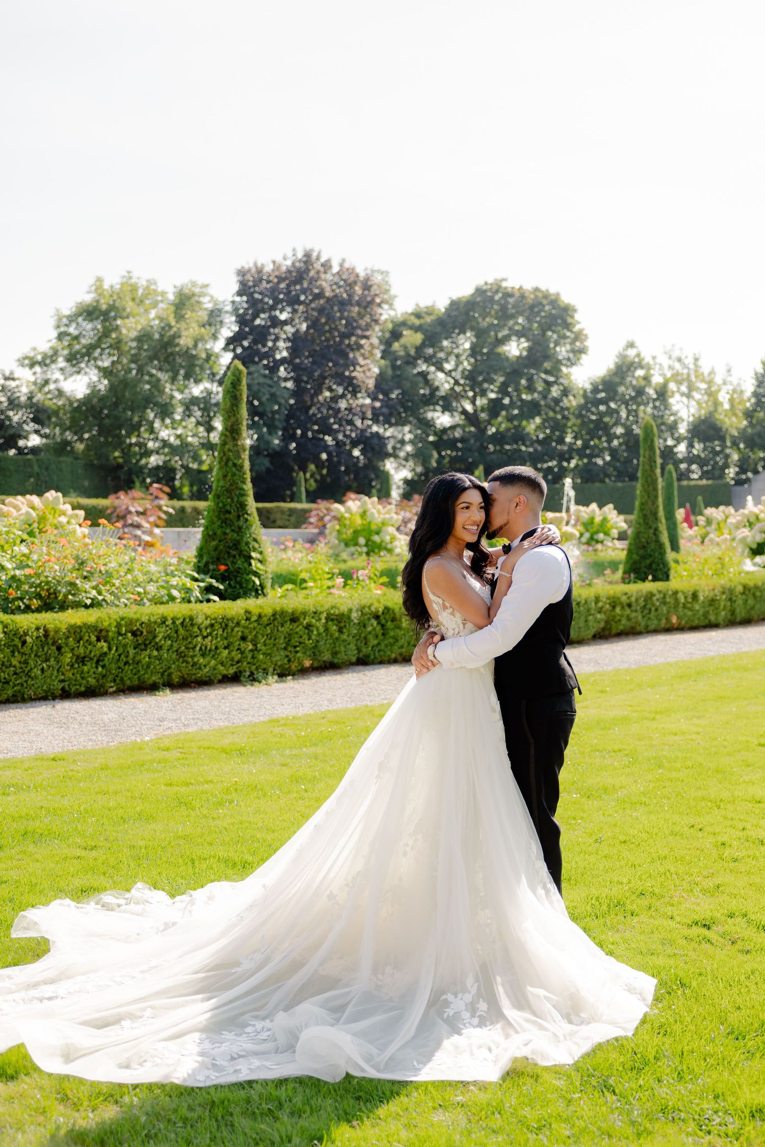 Wedding couple shares a tender moment in a sunlit garden with ornamental trees and flowers.