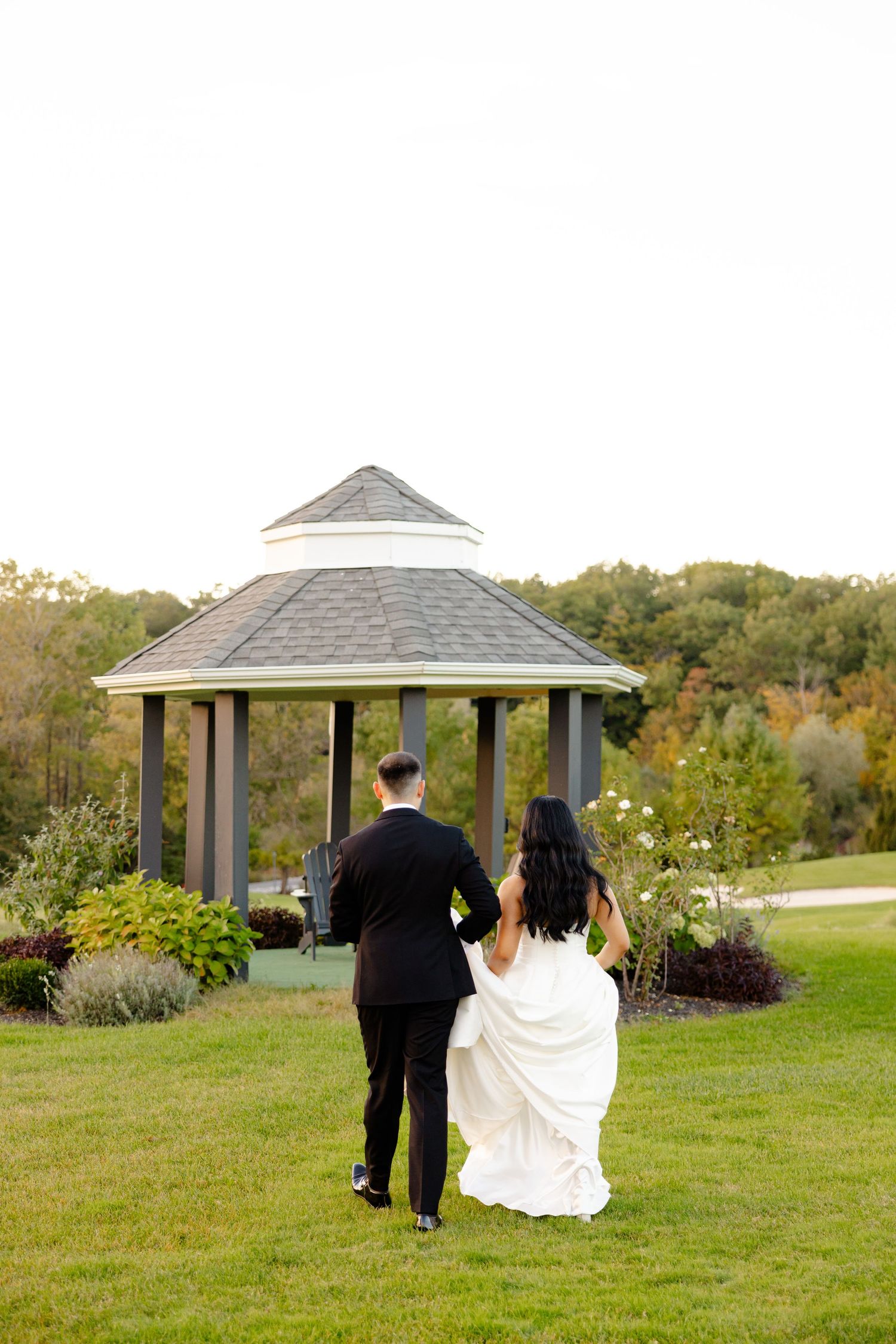 Couple walks towards garden gazebo on their wedding day surrounded by autumn foliage.
