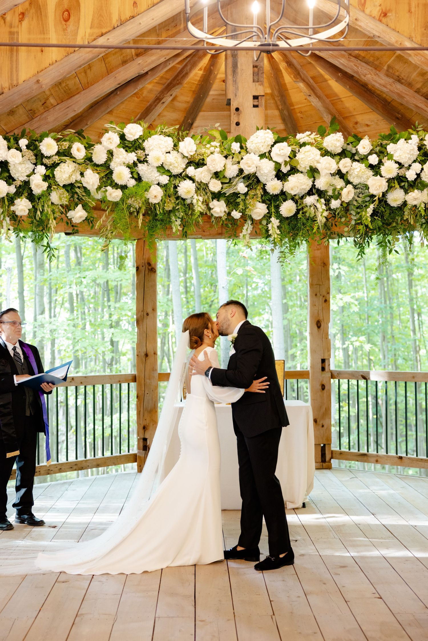 A ceremony takes place in a rustic gazebo adorned with white floral arrangements and natural lighting.