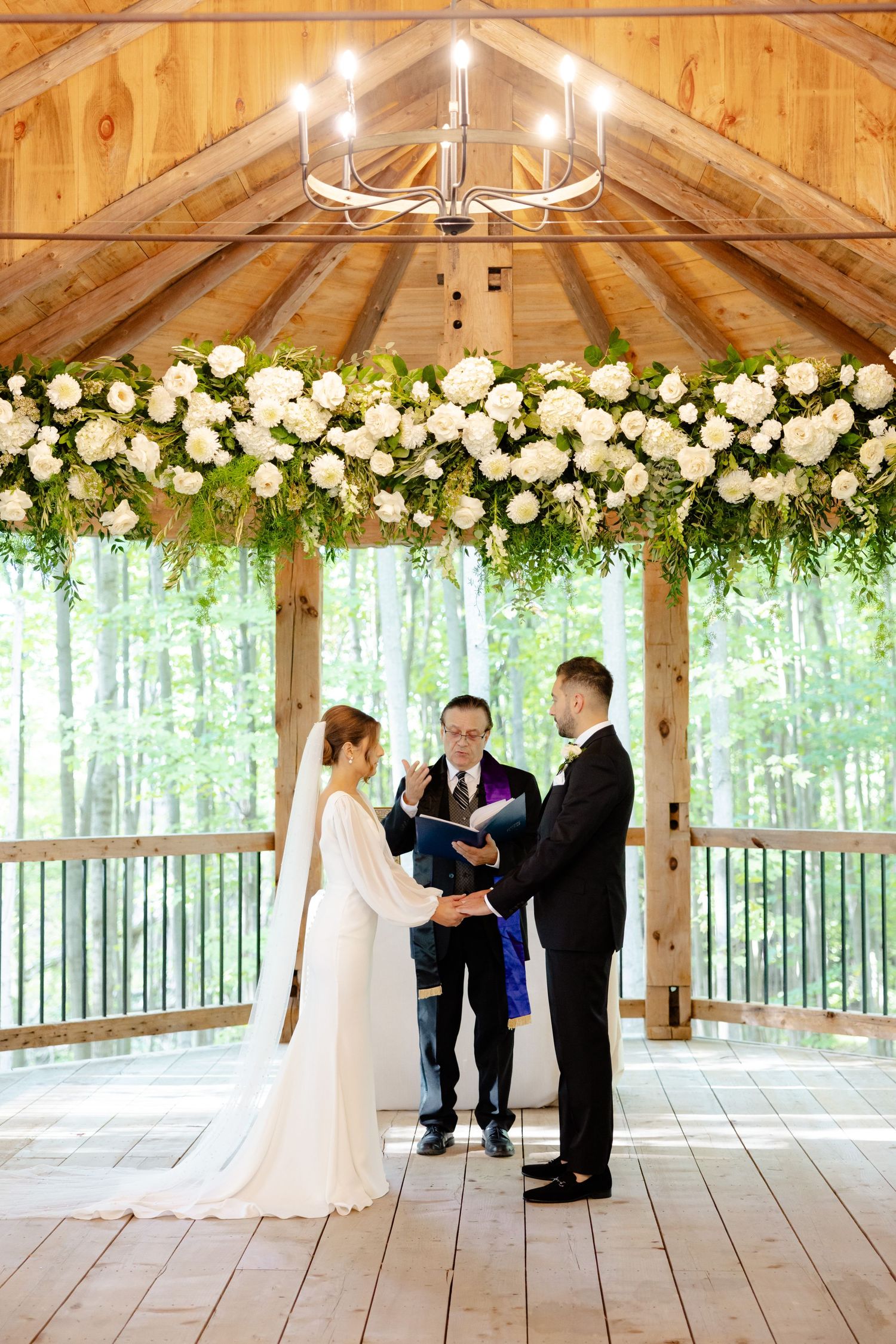 A wedding ceremony beneath an elegant chandelier and floral arch in a rustic wooden pavilion.
