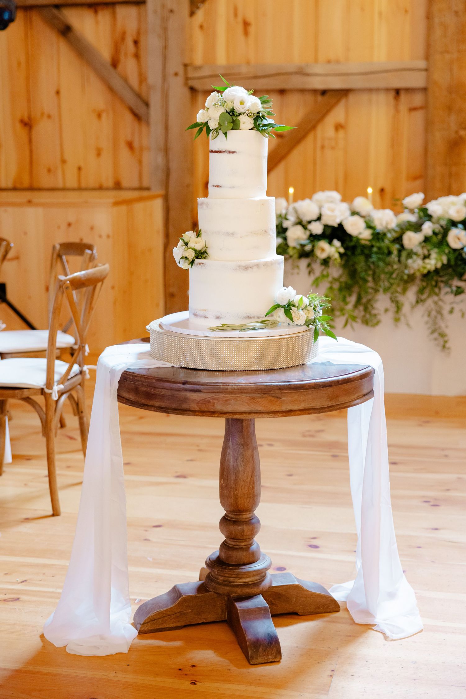 Three-tiered white wedding cake displayed on a rustic wooden pedestal with draped fabric.