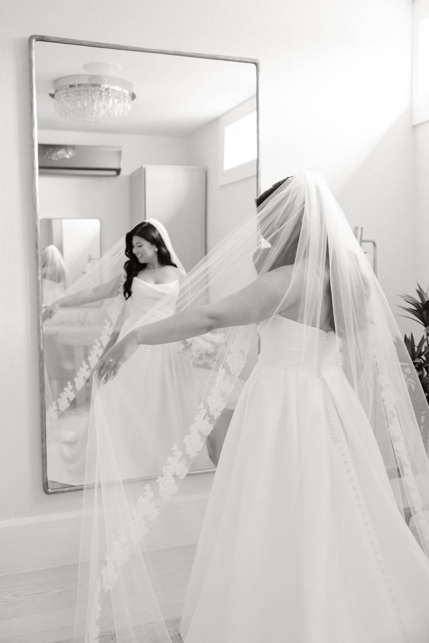 Black and white image of bride with flowing veil posing in front of mirror.