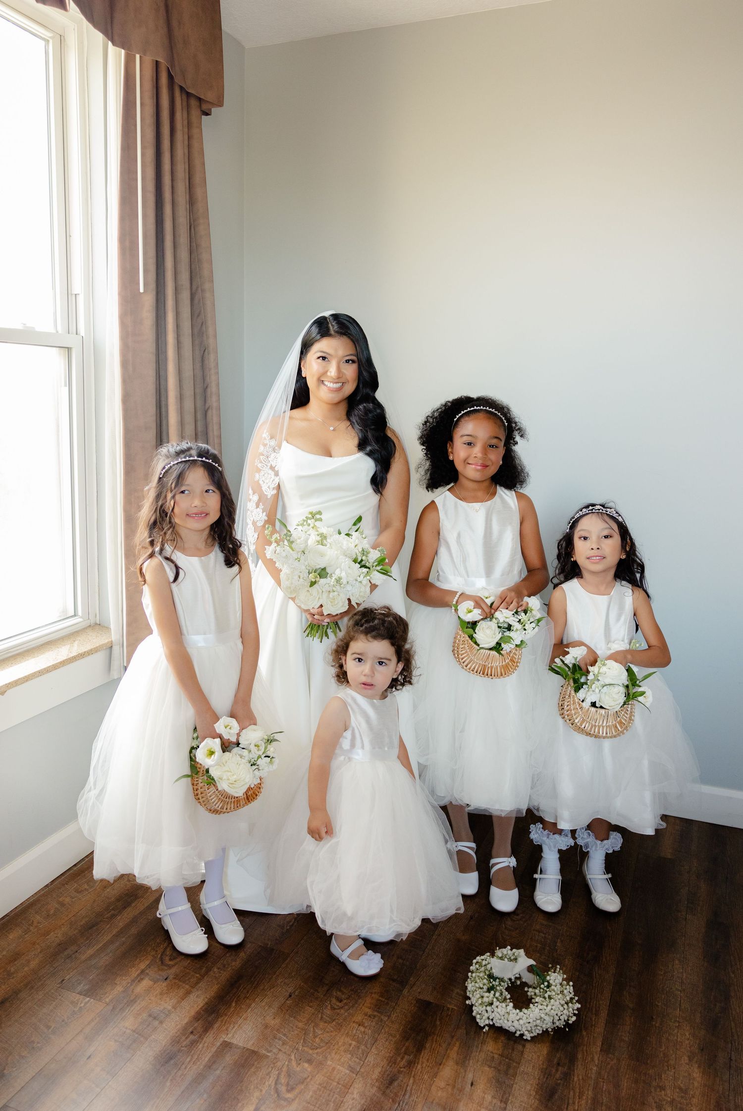 Flower girls in white tulle dresses holding floral baskets pose with the bride.