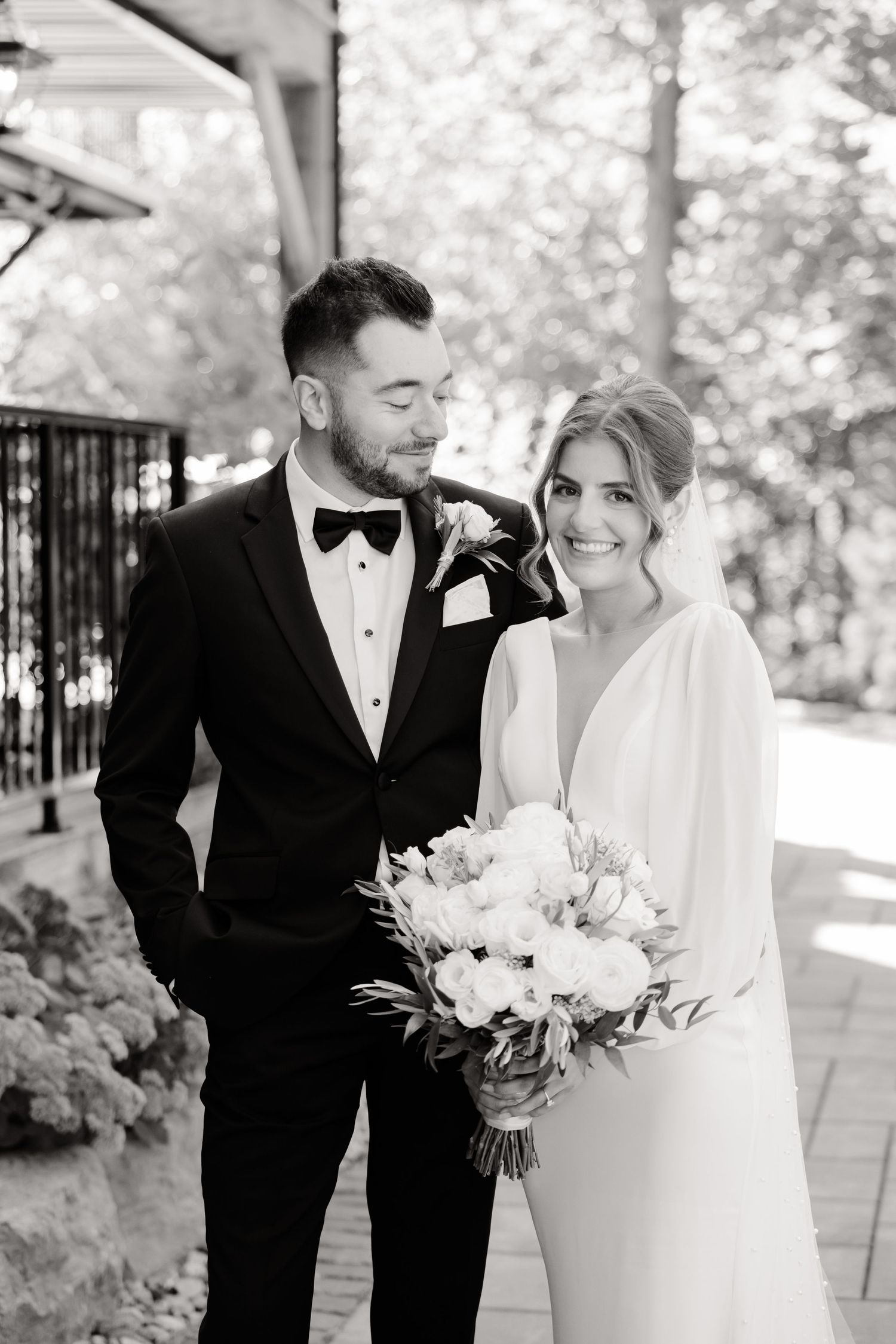 Black and white portrait of newlyweds in classic attire smiling at each other with a white bouquet.
