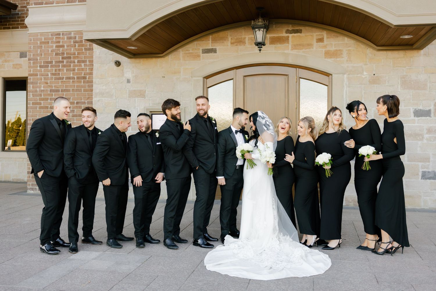 Wedding party in coordinated black attire gathers around bride in white gown for formal portrait.