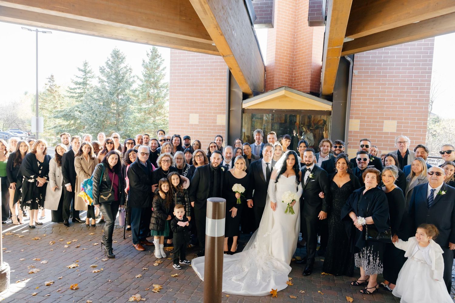 A large wedding party group photo taken outside a modern church building.