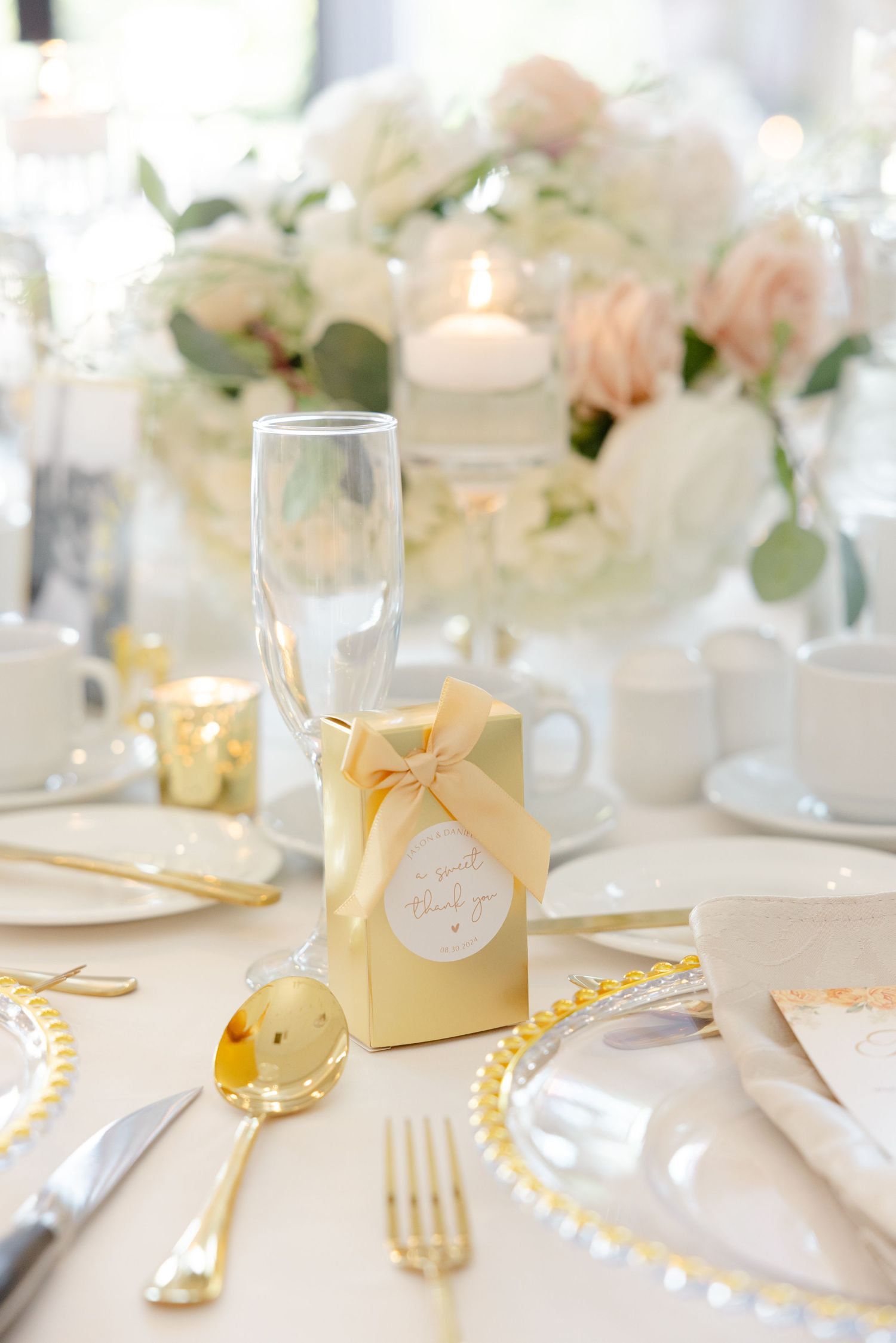 Wedding table place setting with gold favor box, champagne flute, and soft pink floral centerpiece.