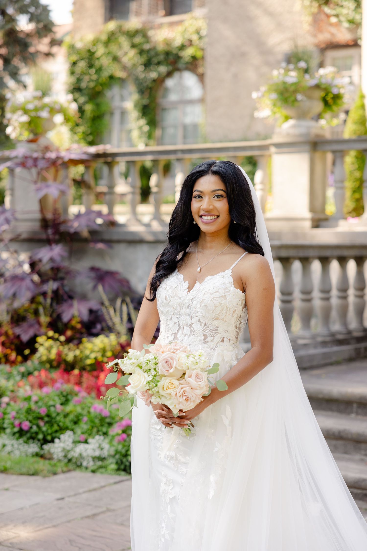 A wedding dress with intricate lace detail and long veil photographed in front of a historic stone building with flowers.