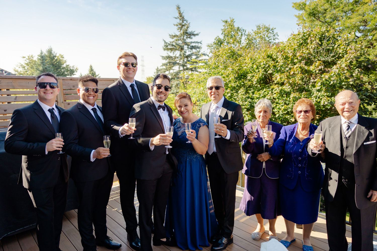 Wedding party in formal attire raising glasses for celebratory toast on outdoor deck.