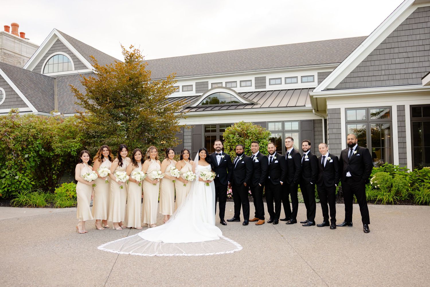 Wedding party in champagne and black attire poses in front of a coastal-style building with autumn foliage.