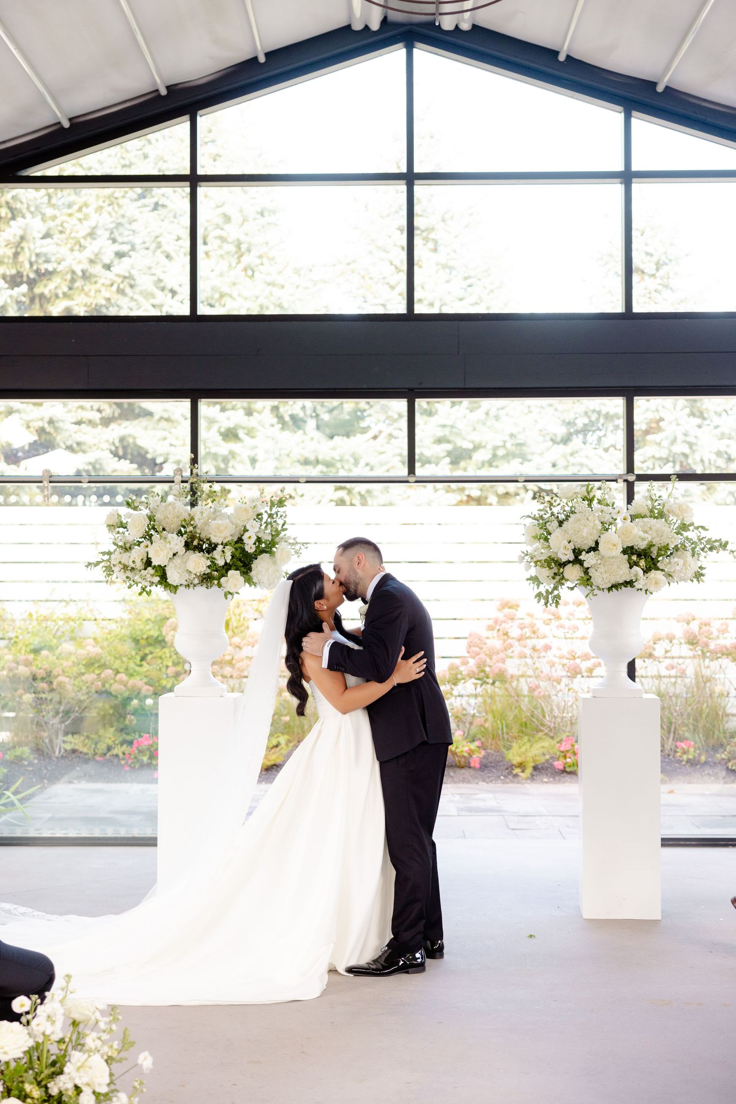 A wedding couple shares a romantic kiss at the altar framed by large windows and white floral arrangements.