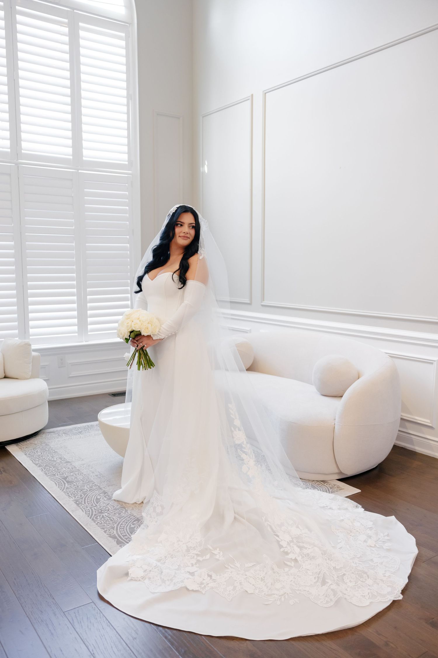 Bride poses in elegant white room with cathedral train and white rose bouquet.