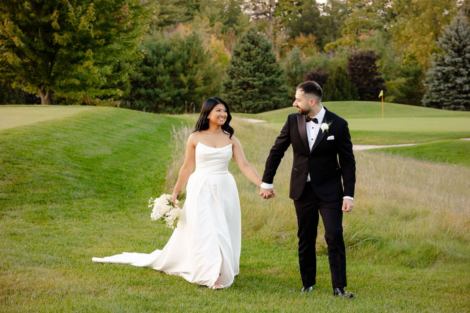A wedding couple walks hand in hand across a manicured lawn with evergreen trees in the background.