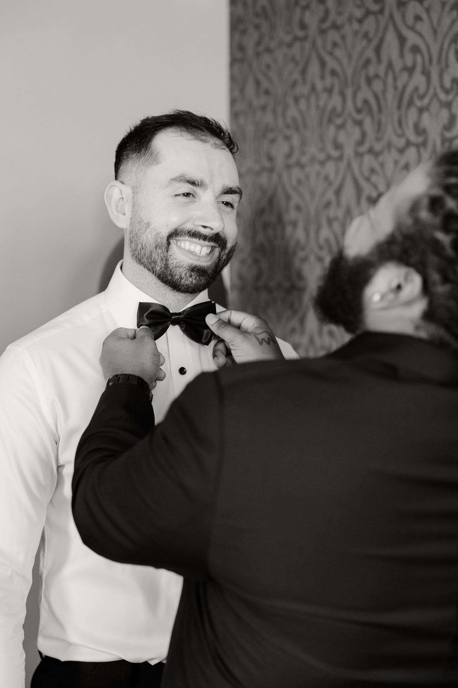A black and white photo of wedding preparations showing a bow tie adjustment moment.
