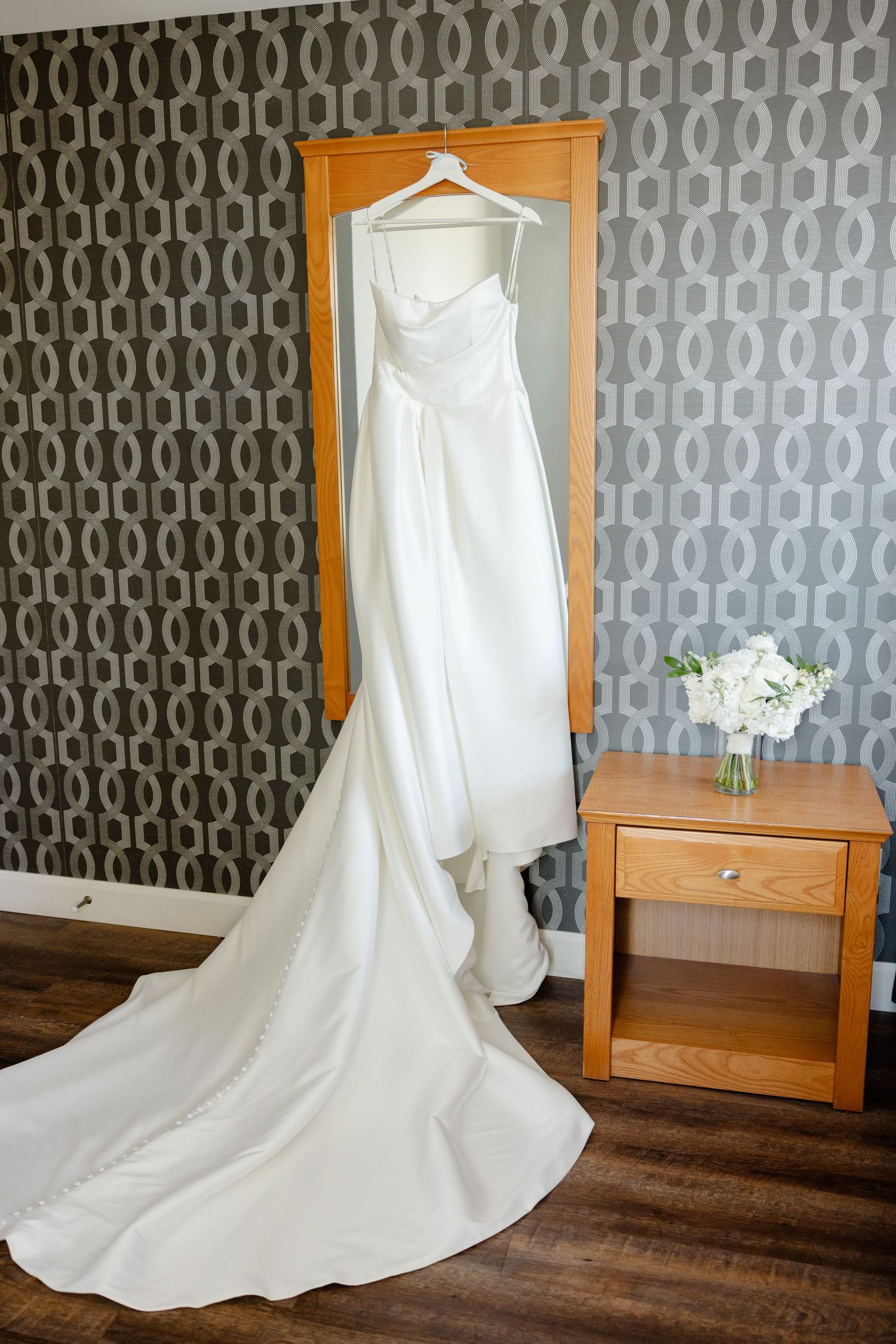 White wedding gown displayed on hanger with flowing train against geometric wallpaper and wooden side table.