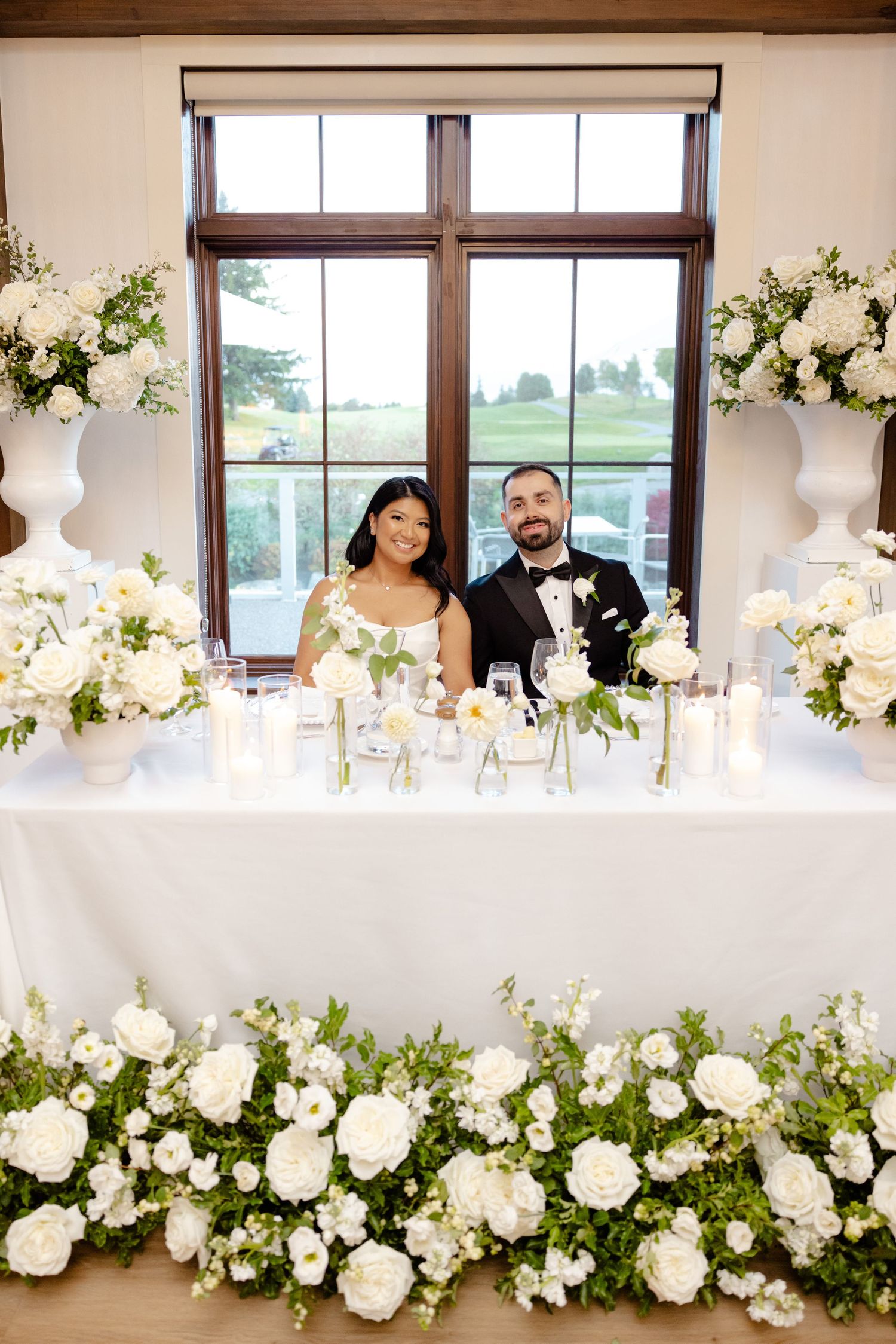 A sweetheart table adorned with lush white floral arrangements in front of a large window.