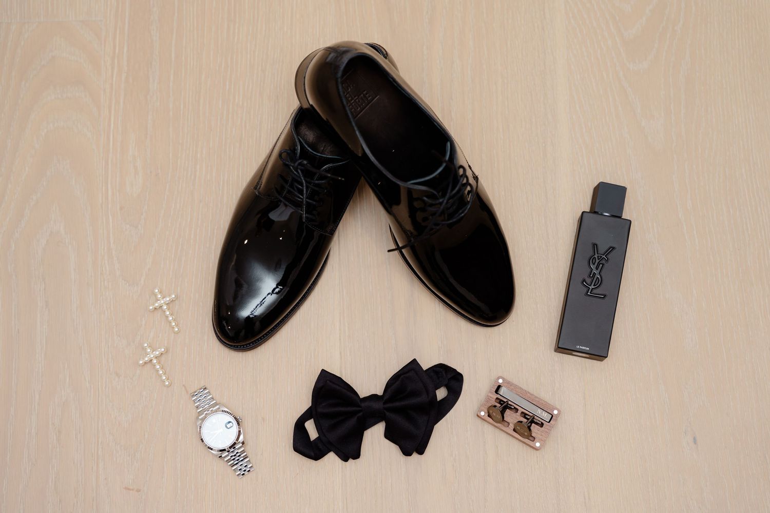Wedding accessories including black dress shoes, bowtie, watch and USB drive on wooden floor.