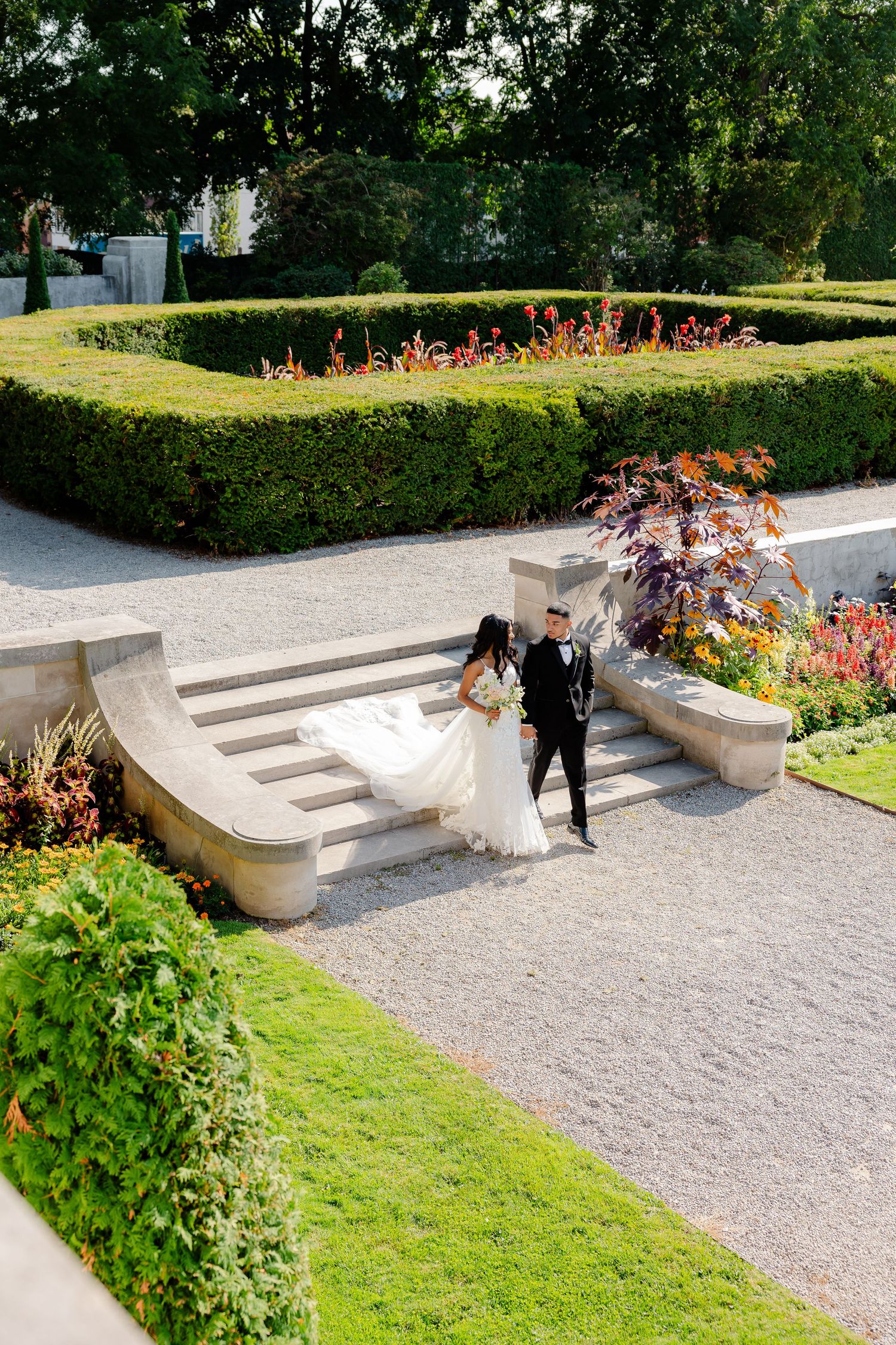 Wedding couple descending elegant garden steps surrounded by colorful flower beds and hedges.
