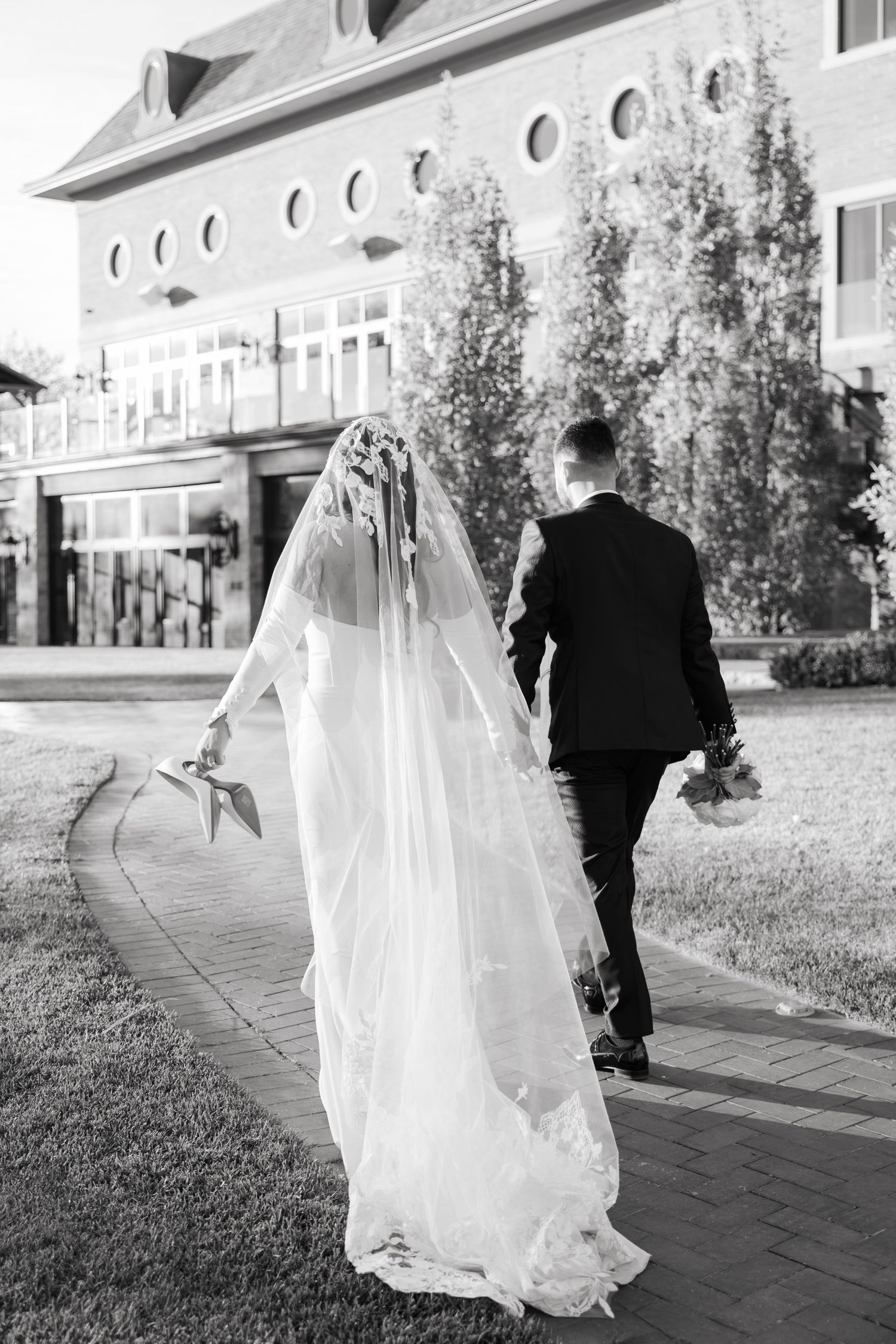 Black and white photo of bridal couple walking away on brick path near historic building.