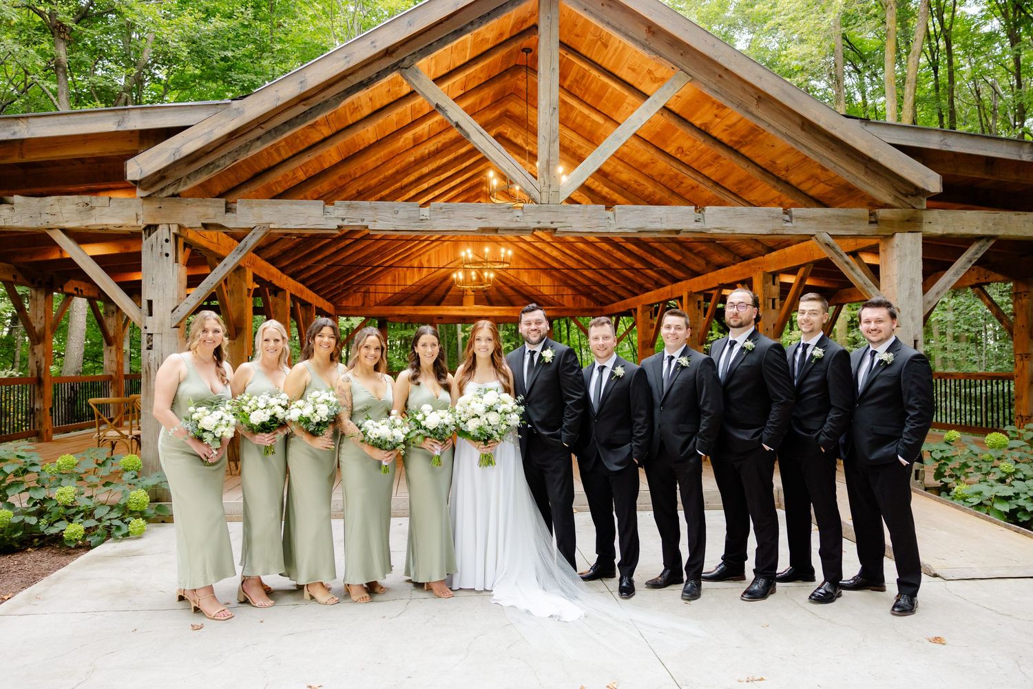 Wedding party members in black suits and champagne dresses pose under wooden pavilion roof.