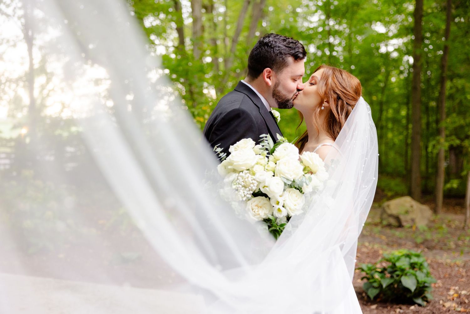 Romantic wedding kiss captured through flowing veil with white roses in natural setting.