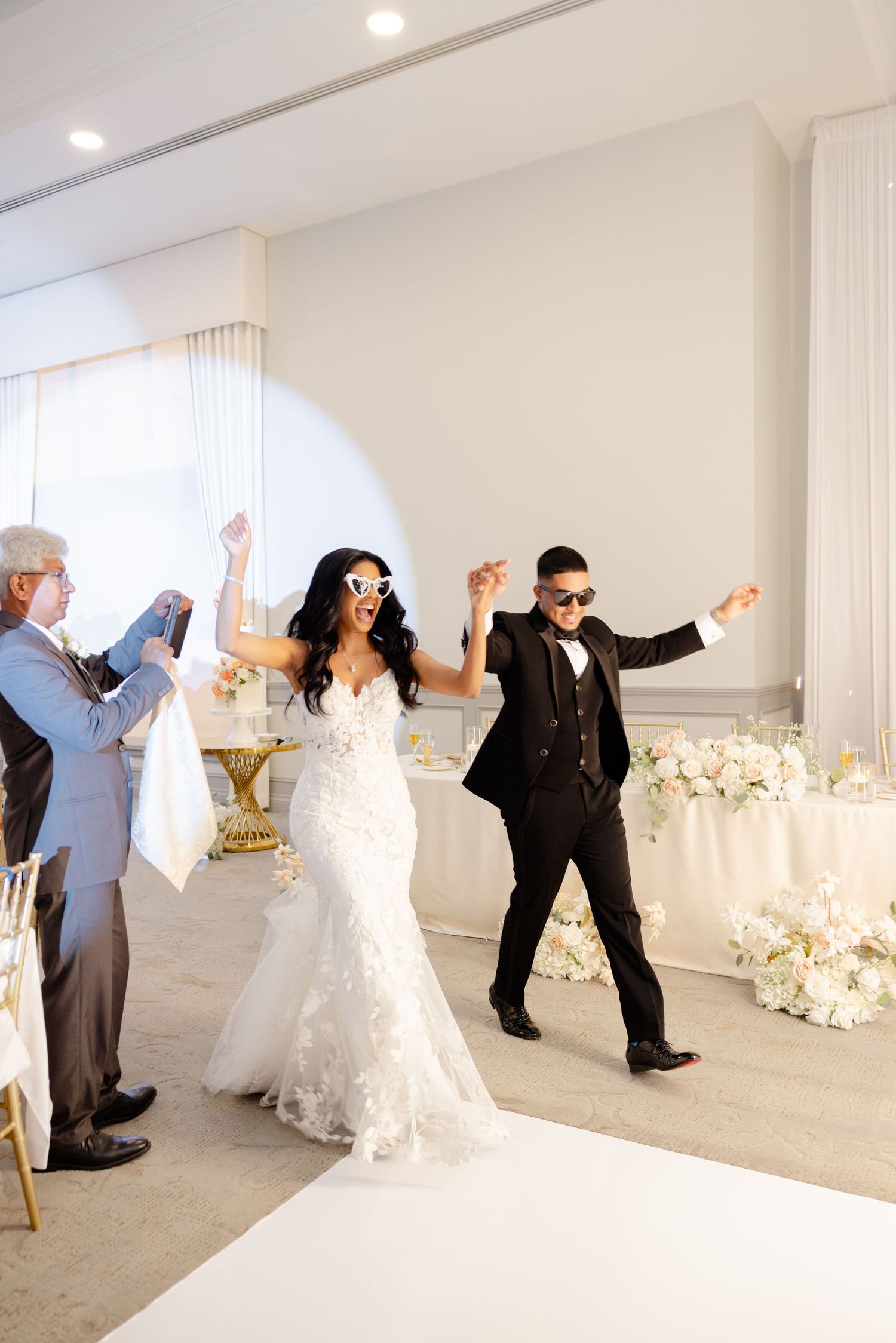 A couple celebrates during their wedding reception with joyful dance moves in an elegant white venue decorated with flowers.