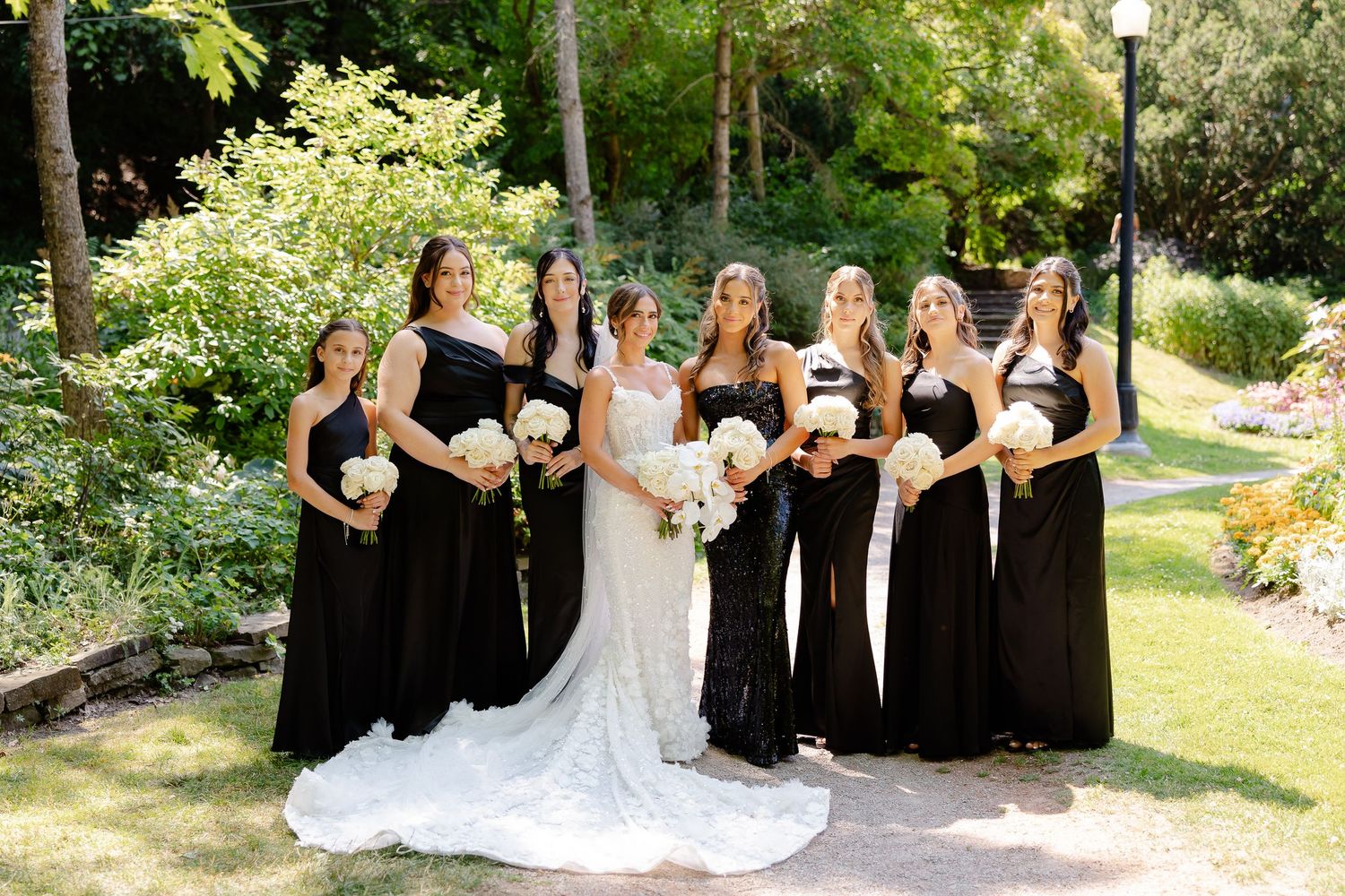 Wedding party poses outdoors with bridesmaids in black gowns and bride in white lace dress holding bouquets in garden setting.