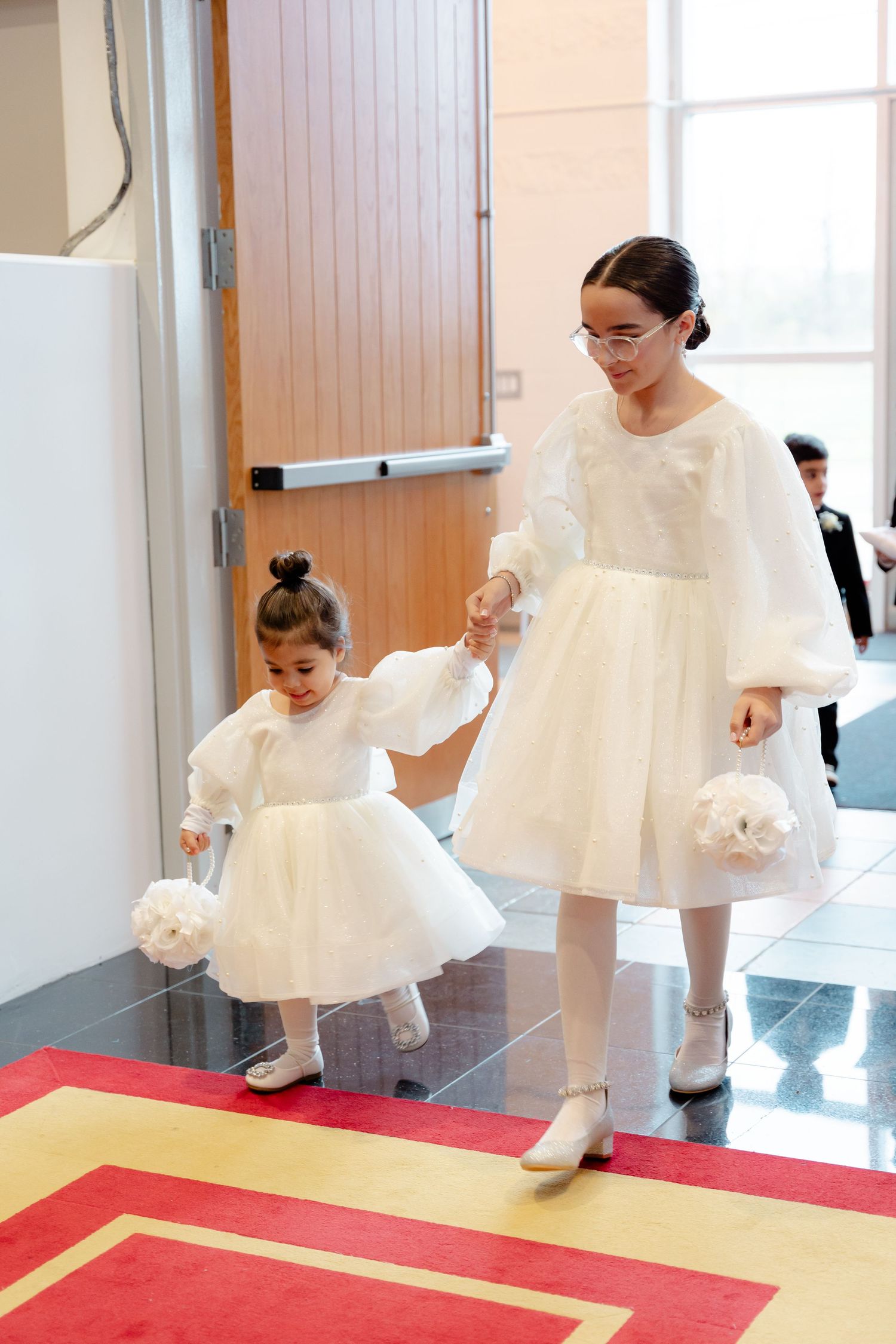 Woman in white dress holds hands with young girl in matching dress walking down red carpet.