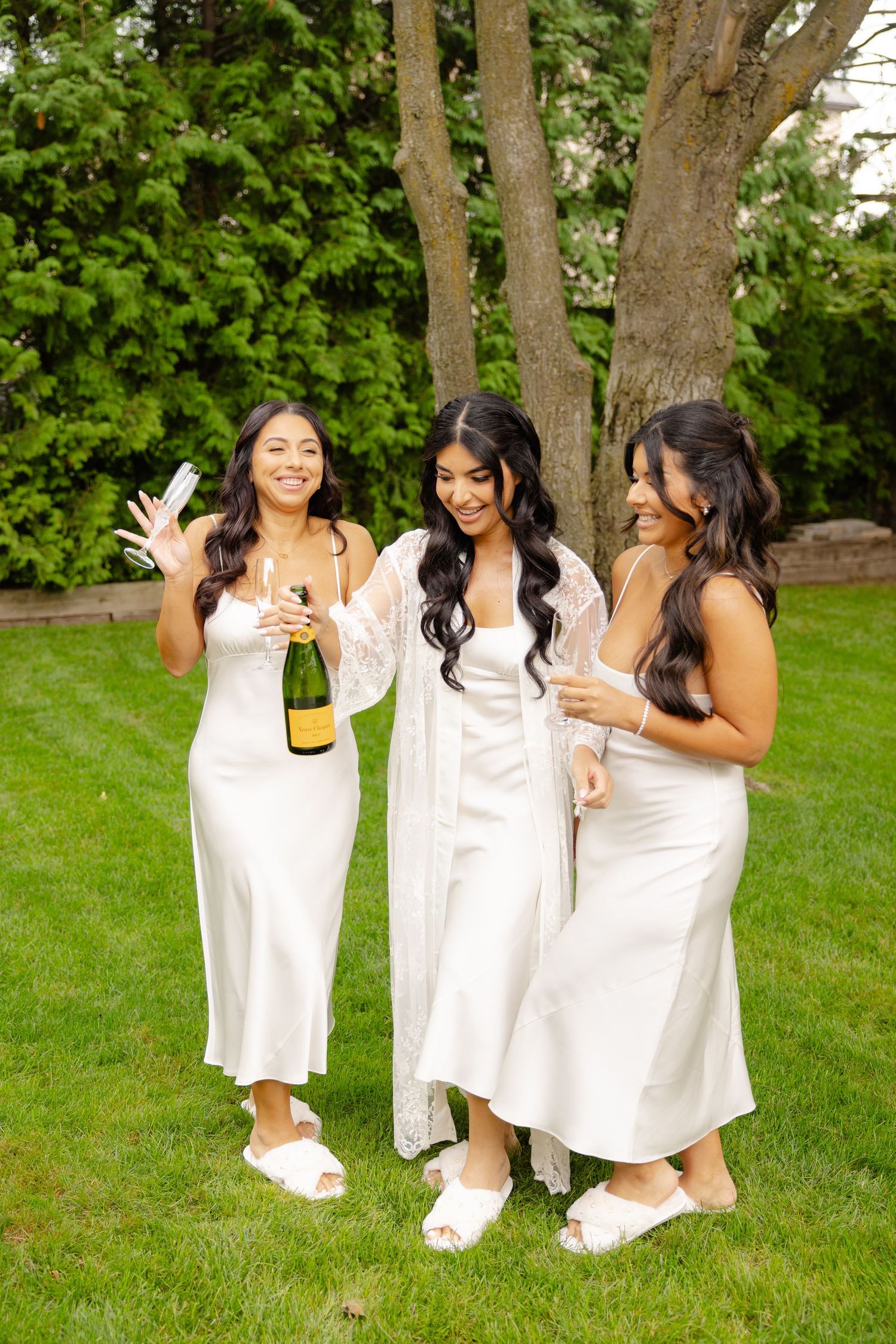 Three women in white dresses laughing and celebrating outdoors on a grassy lawn with trees in background.