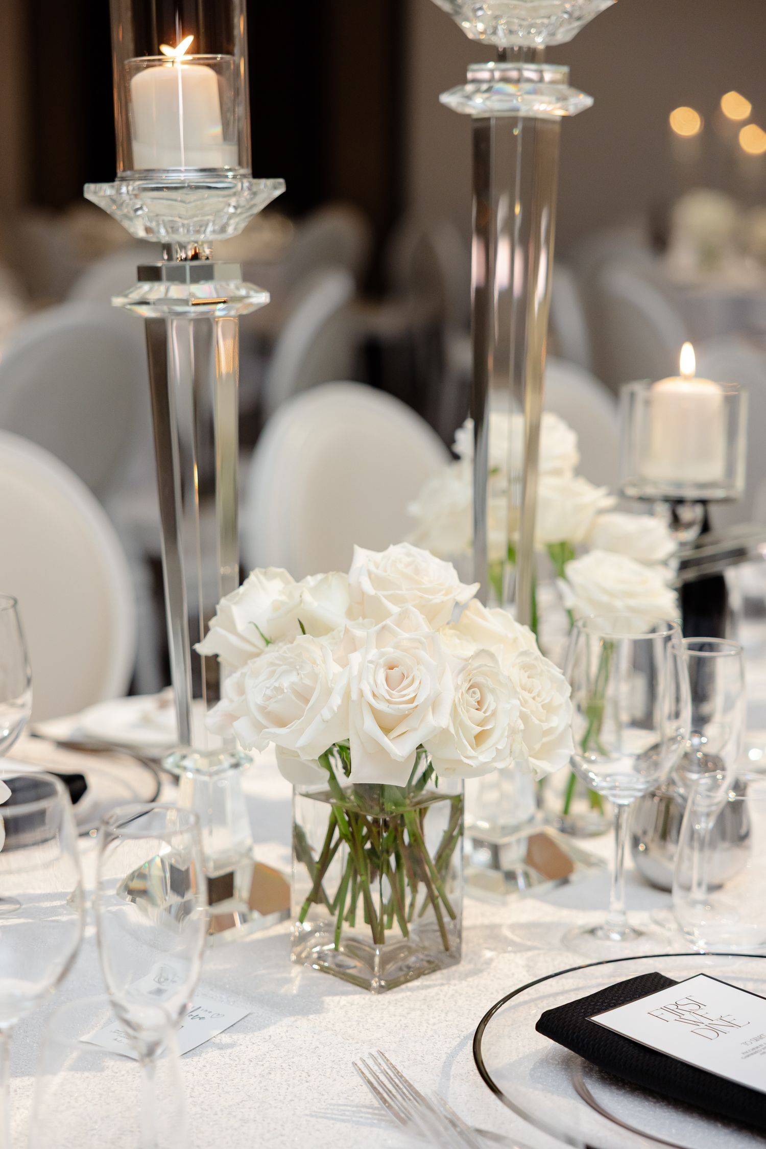 Elegant wedding reception table setting with white roses, crystal candleholders and glowing candles in a formal dining room.