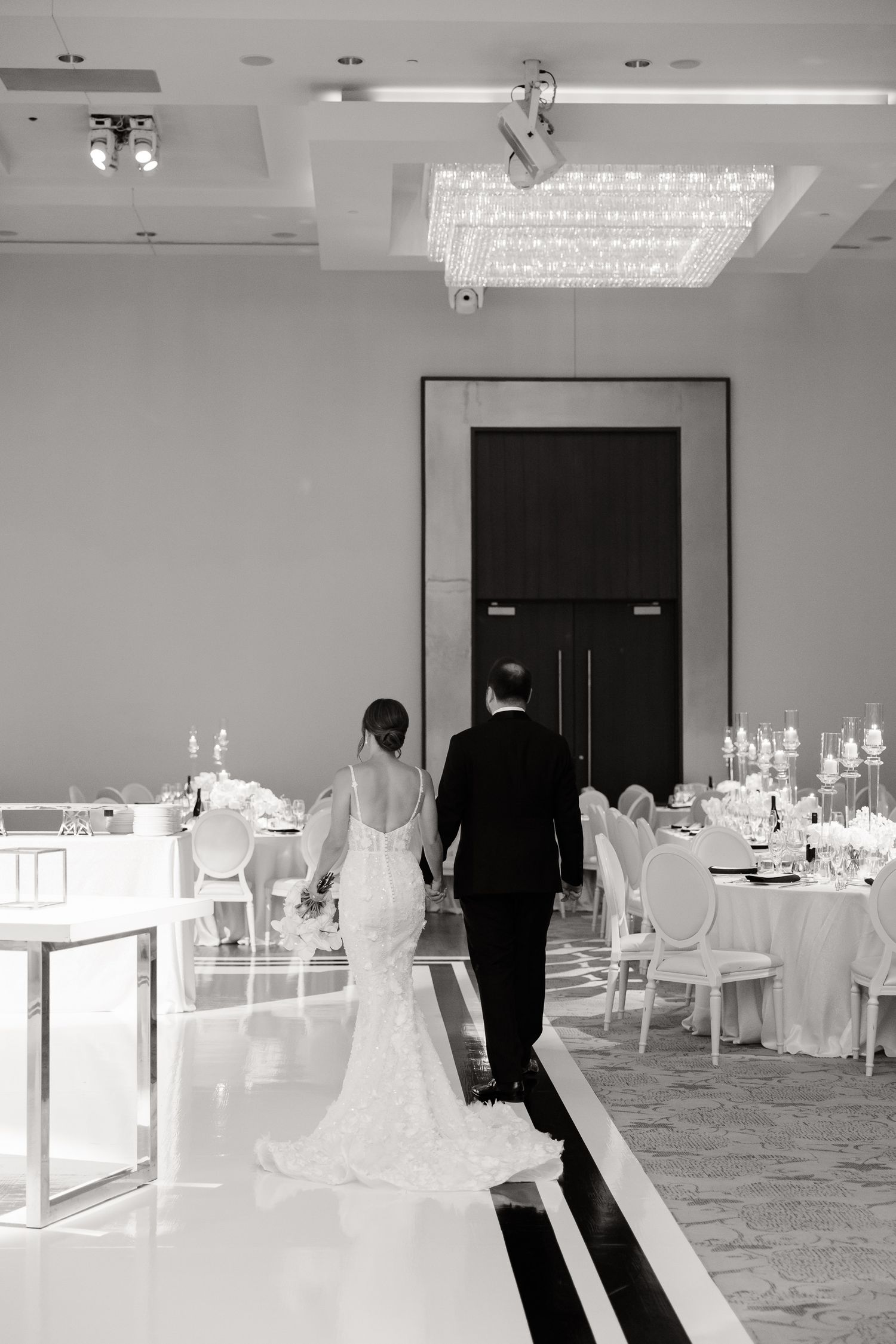 A couple in formal wedding attire walk down a black-and-white striped aisle in an elegant reception hall with crystal lighting.