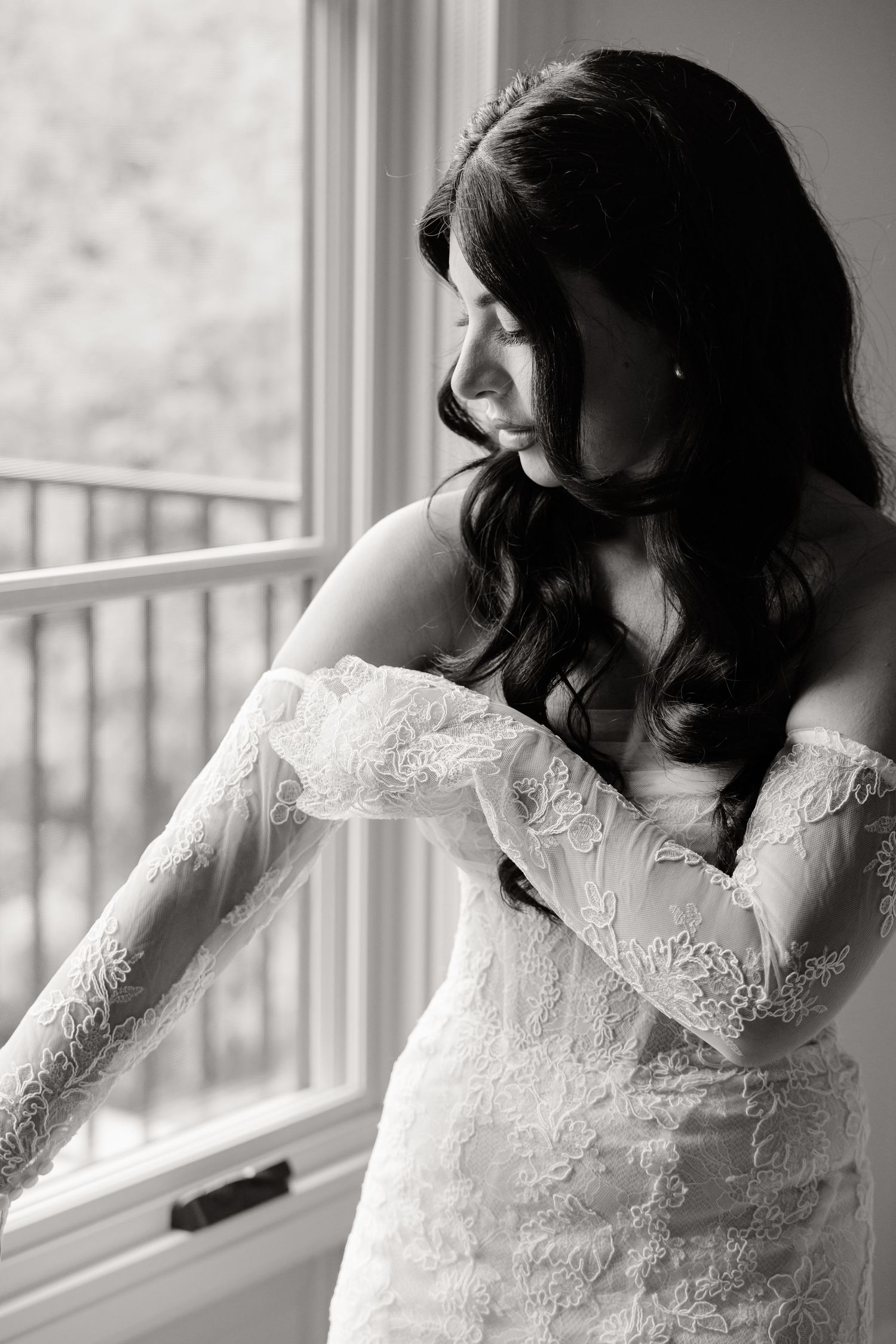 Woman in white lace dress gazes pensively through bright window in black and white photograph.