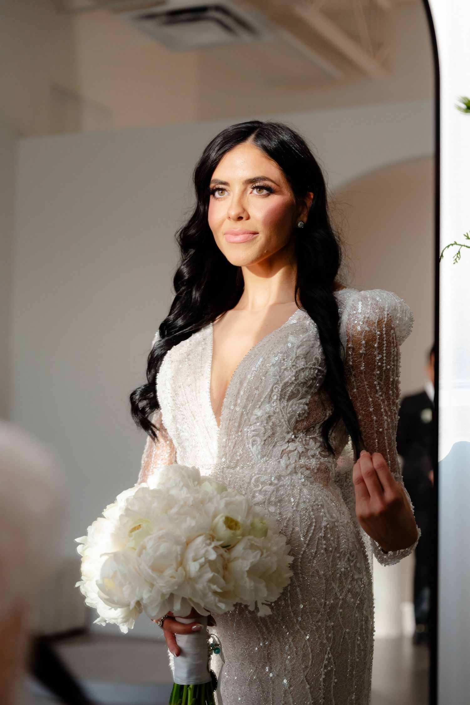 A bride poses with white flowers in an embellished long-sleeve gown near an arched doorway.