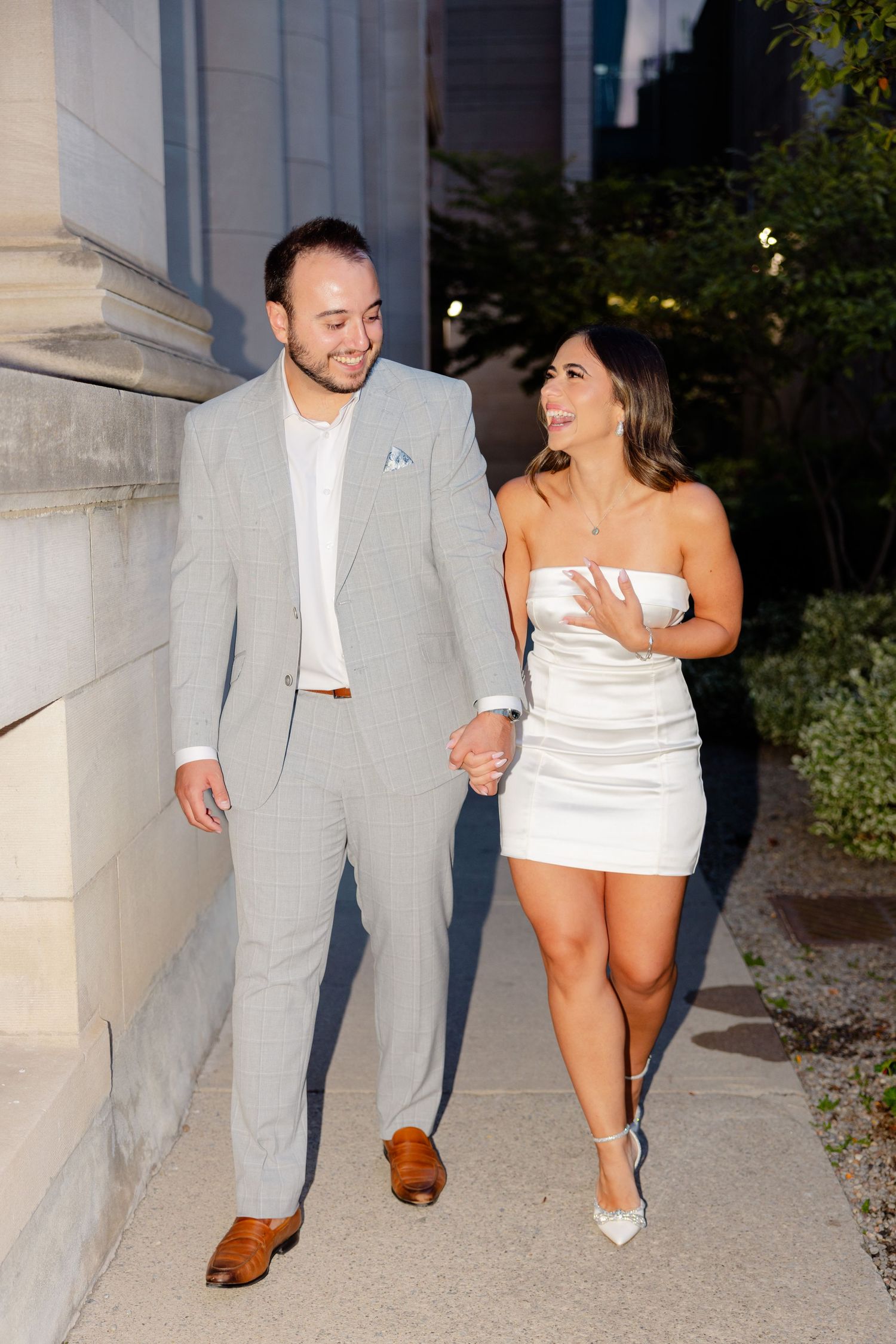 Happy couple walking and laughing together along building exterior in formal wear.