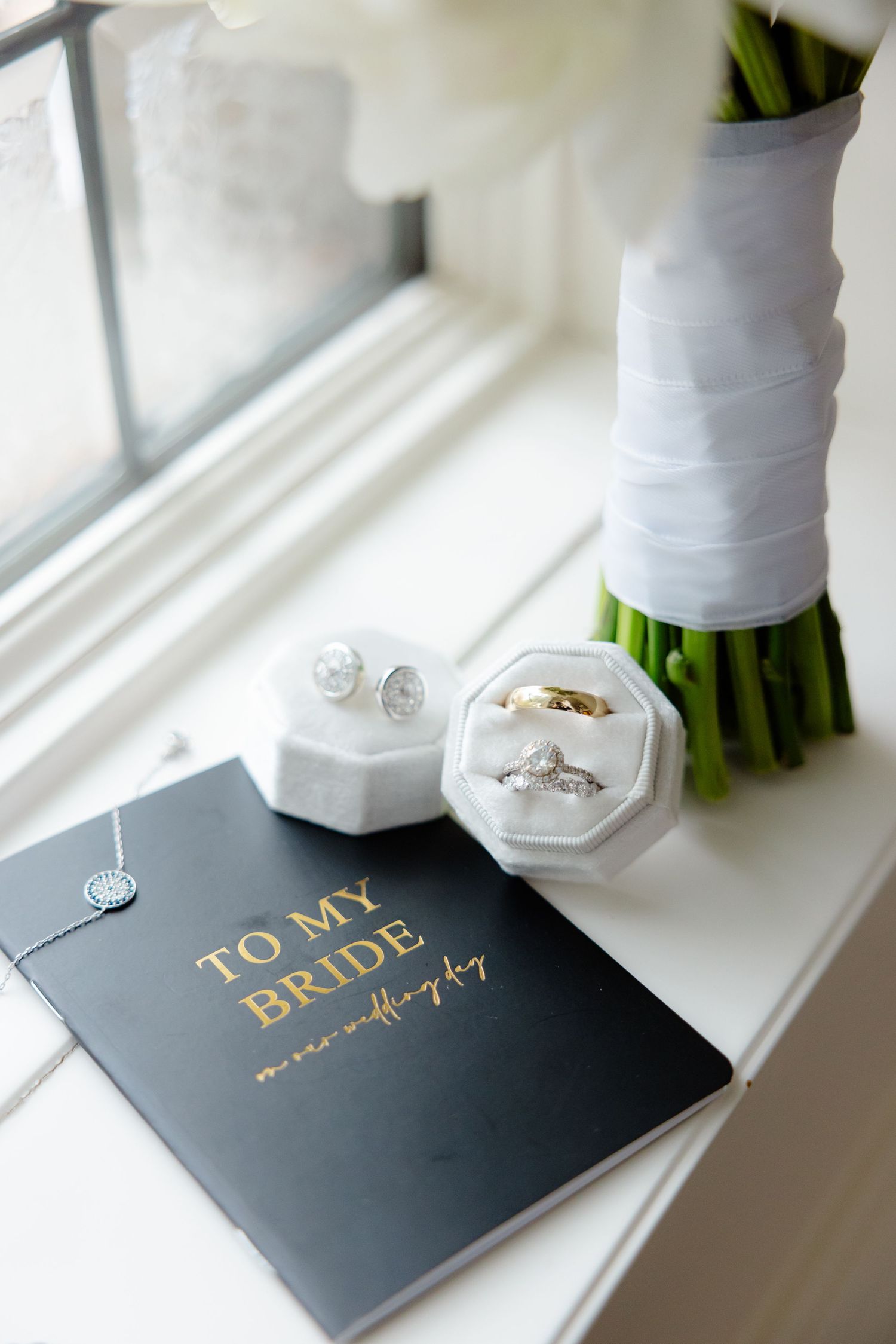Wedding rings and 'To My Bride' book displayed with white bouquet on window ledge.