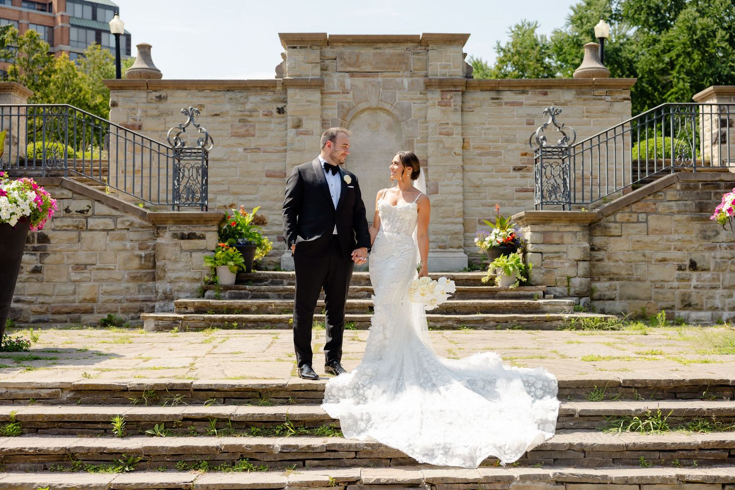 A bride in an elegant lace gown and groom in black suit walk down stone steps at an ornate outdoor venue.