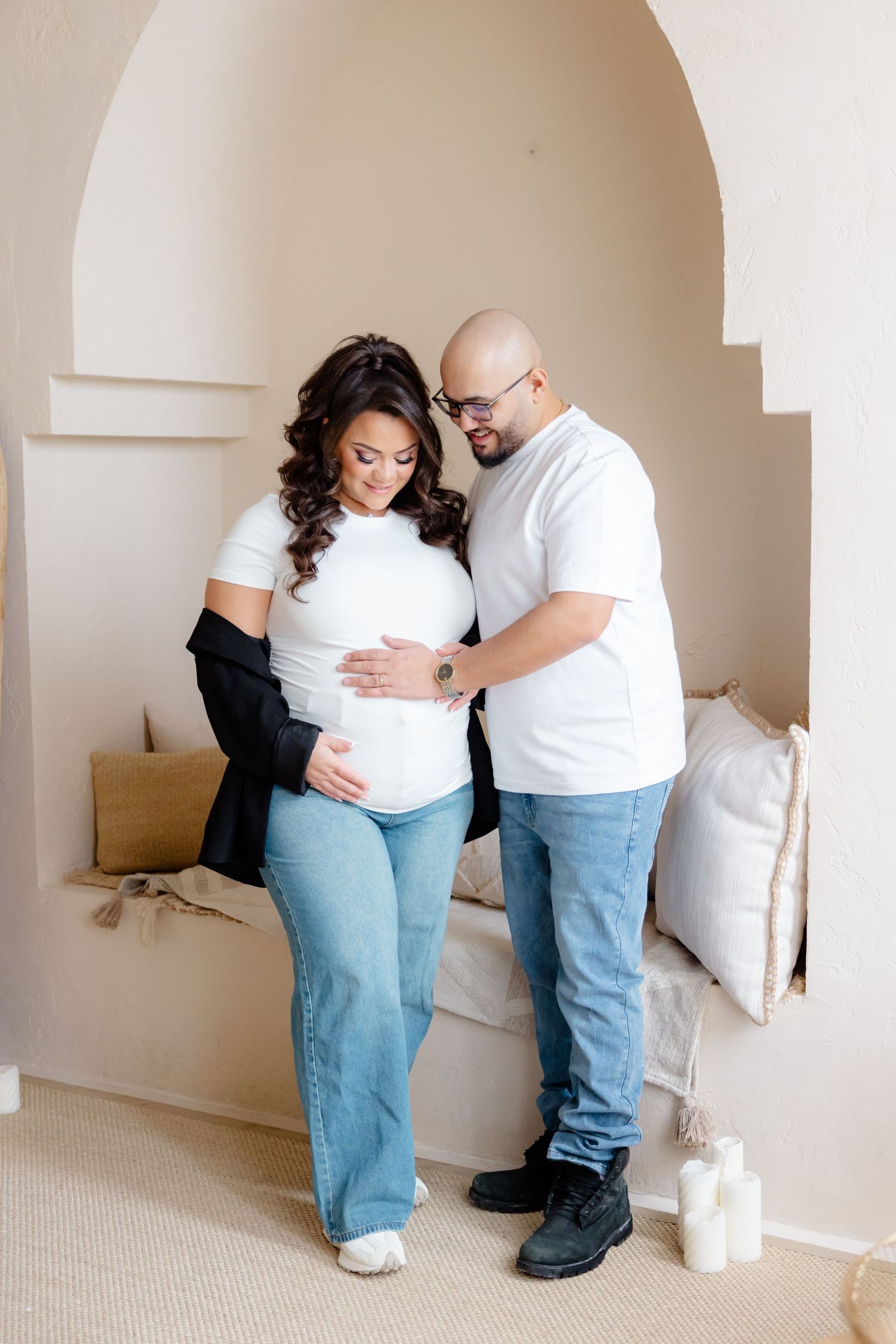 Couple wearing white shirts and blue jeans embrace while standing in an archway at home during a maternity photoshoot.