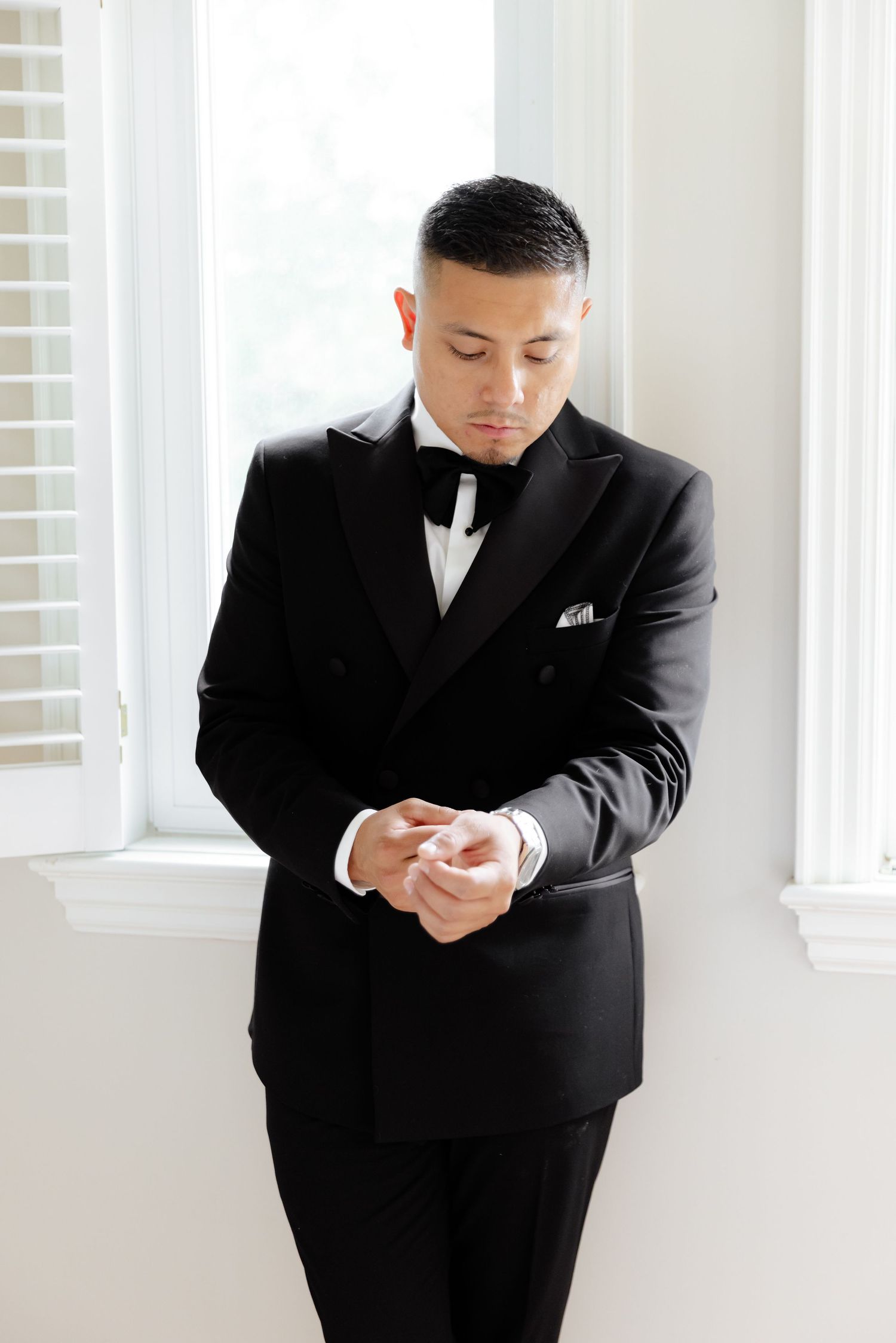A person in a formal black tuxedo looks down at something in their hands by a window with white drapes.