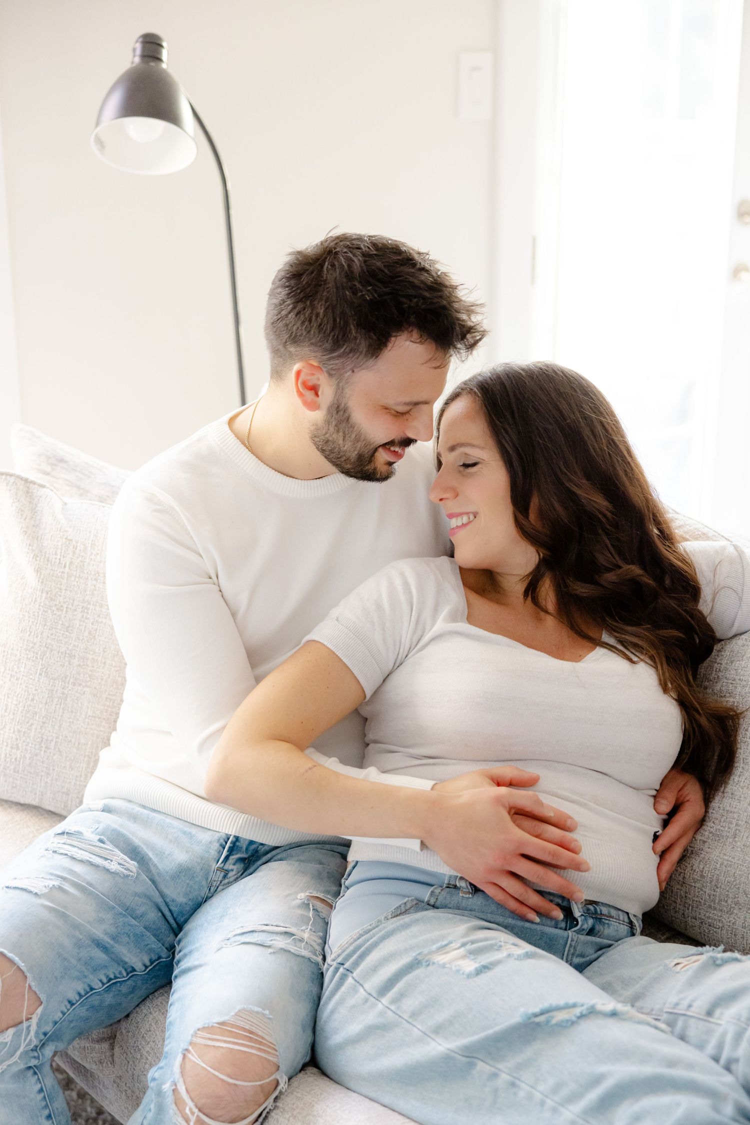 A couple in white shirts and jeans cuddle intimately on a couch, with the woman's pregnant belly visible.