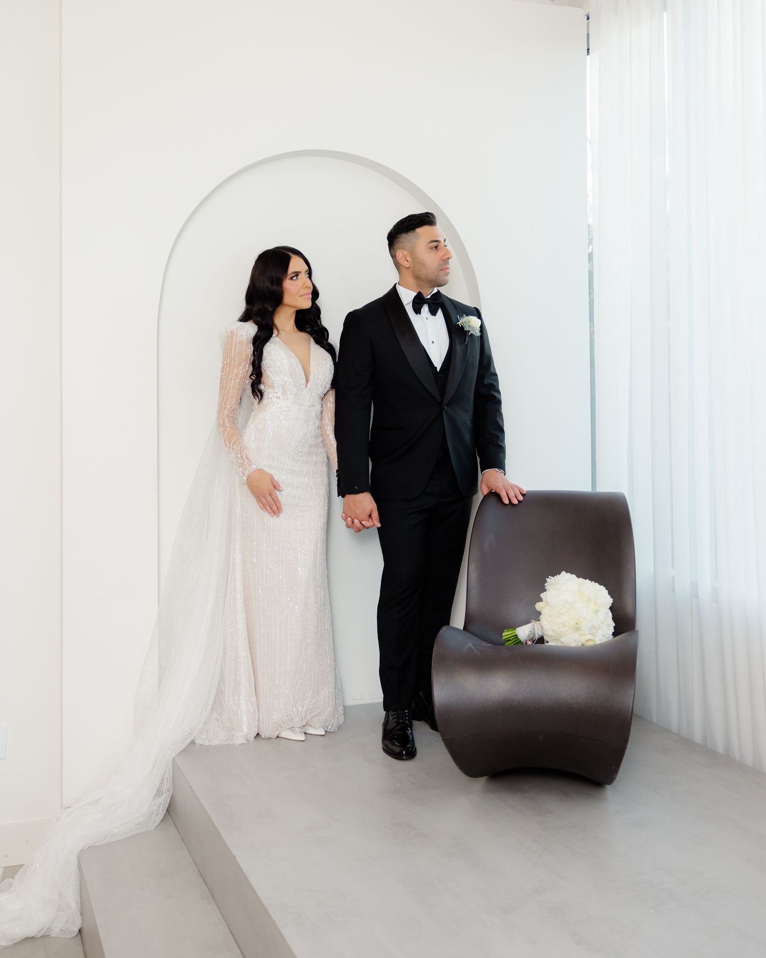 Bride in lace gown and groom in tuxedo posing by modern brown chair with white roses.